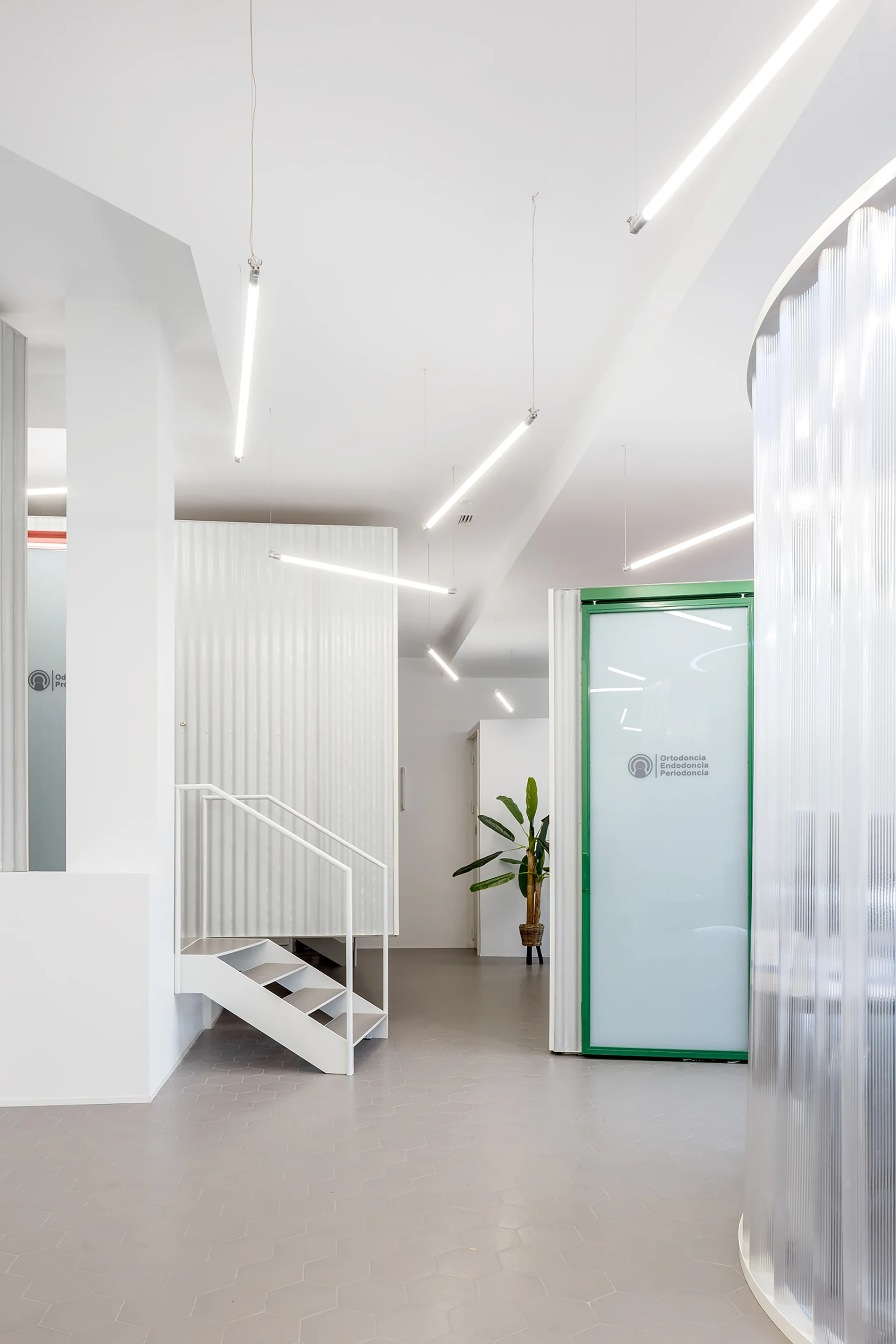 A bright minimalist interior featuring grey hexagonal floor tiles, suspended linear tube lights, a white metal staircase, and a green-framed glass door.