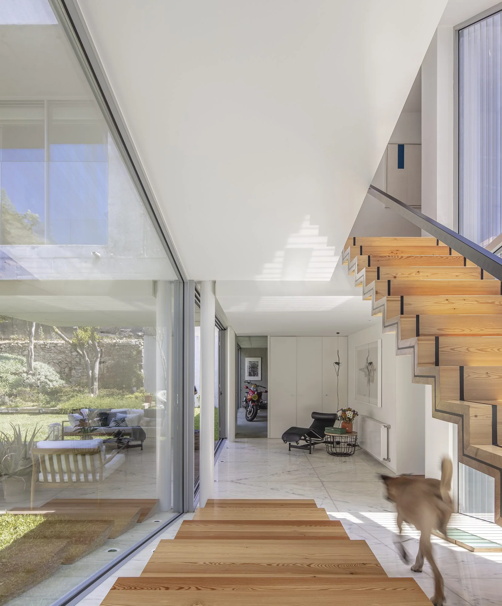 A modern home interior features a light wood open riser staircase next to large glass walls looking out onto a grassy courtyard.