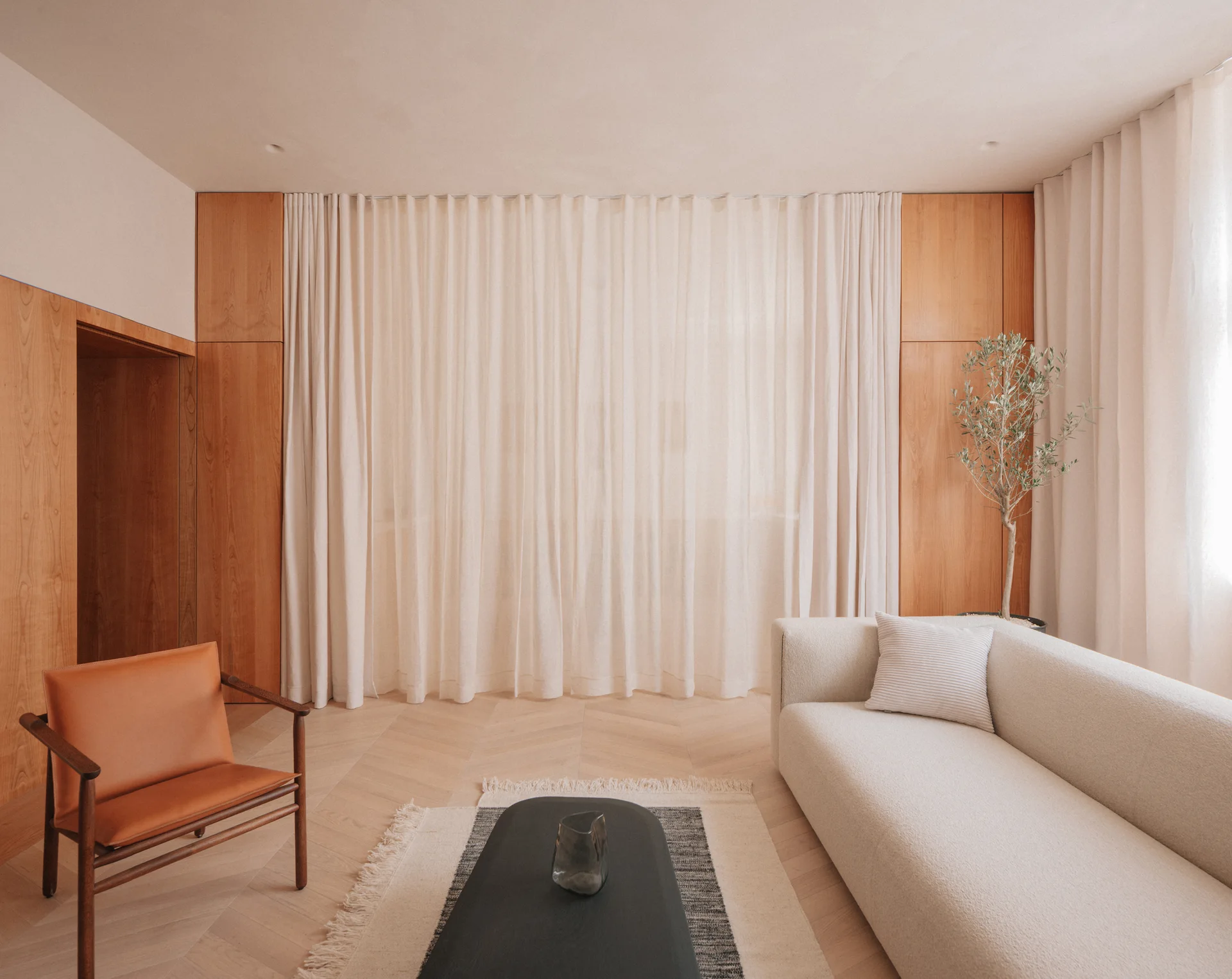 A contemporary living room featuring a cream curved sofa, a tan leather armchair, and floor-to-ceiling sheer curtains set between wood wall panels.