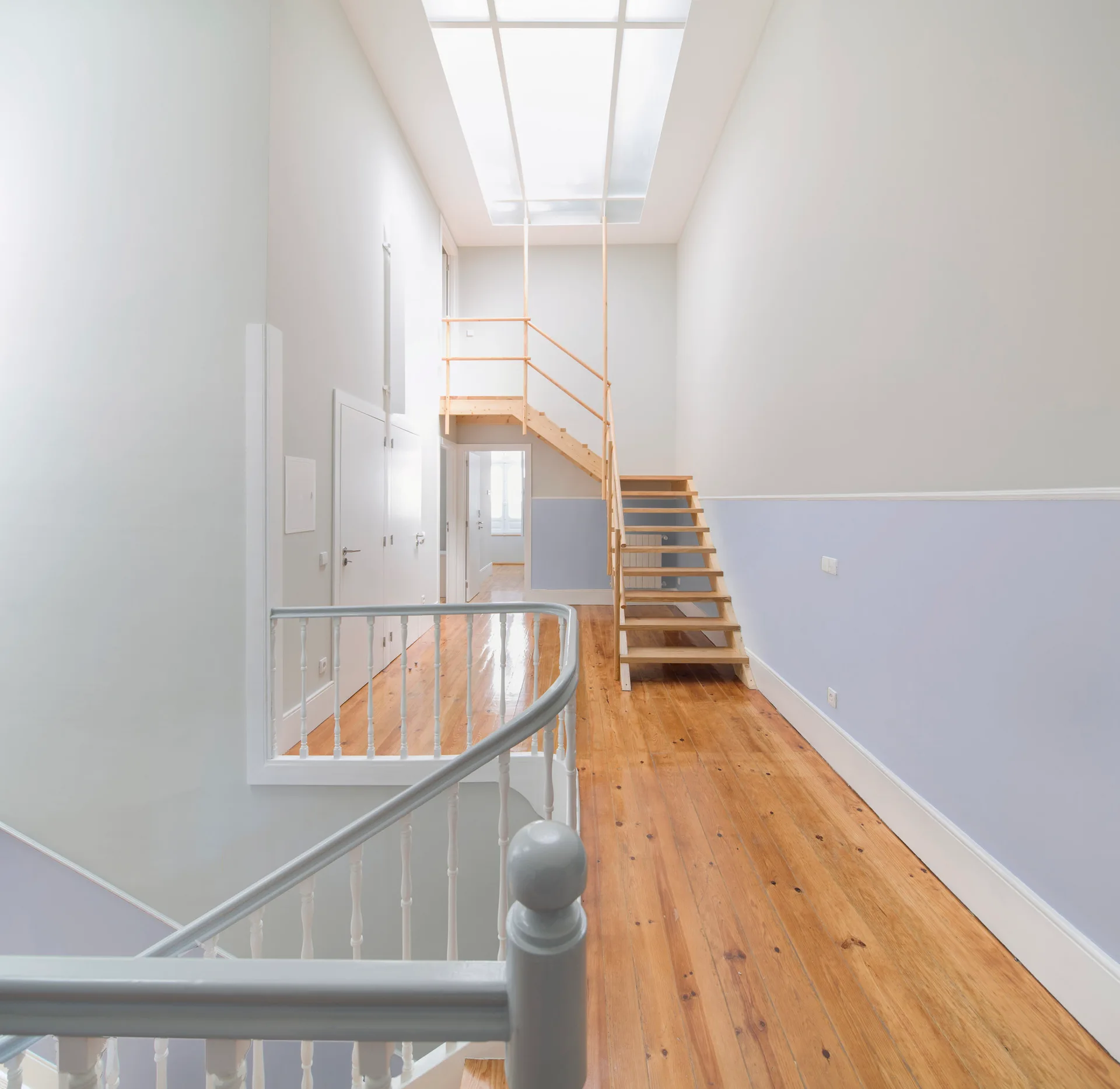 A light-filled hallway features a natural wood plank floor, white and light blue painted walls, an open-riser wooden staircase, and a large skylight.