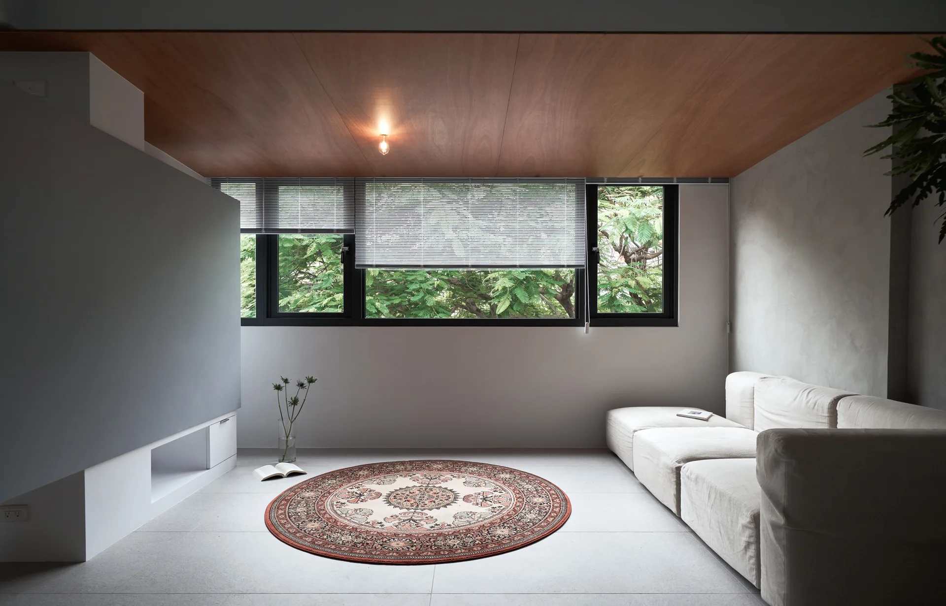 A minimalist living room features a cream modular sofa, a round patterned rug, and a warm wood-paneled ceiling above black-framed windows.