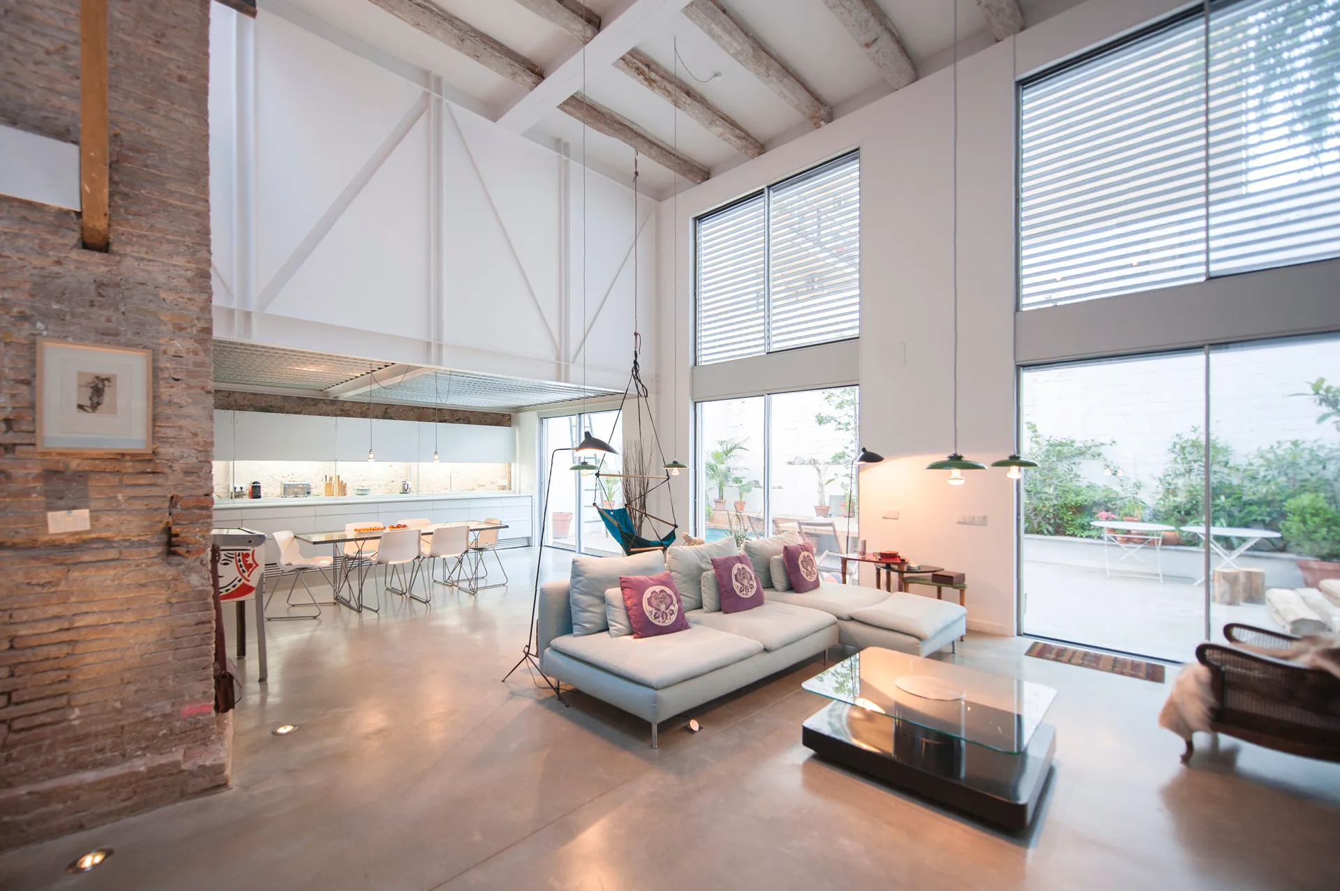 An open-plan industrial loft living room with a modular sectional sofa, polished concrete floor, and exposed ceiling beams next to large glass patio doors.