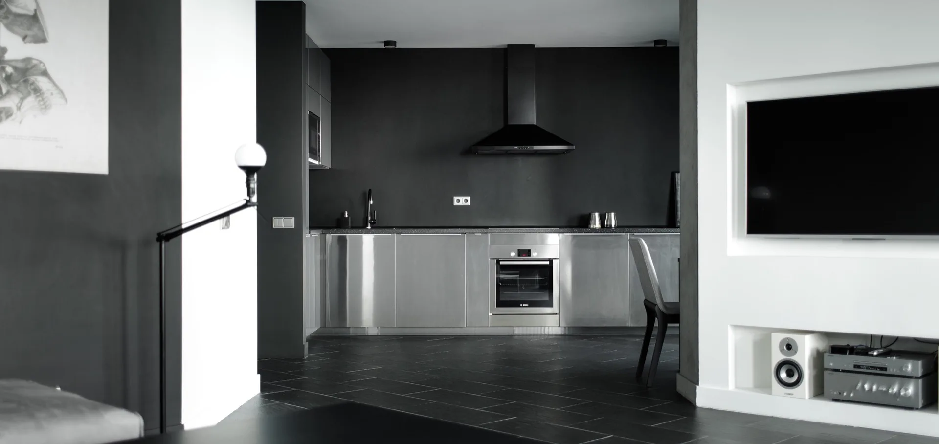 A minimalist kitchen features brushed stainless steel base cabinetry, a matte black wall with a dark range hood, and dark tiled flooring.