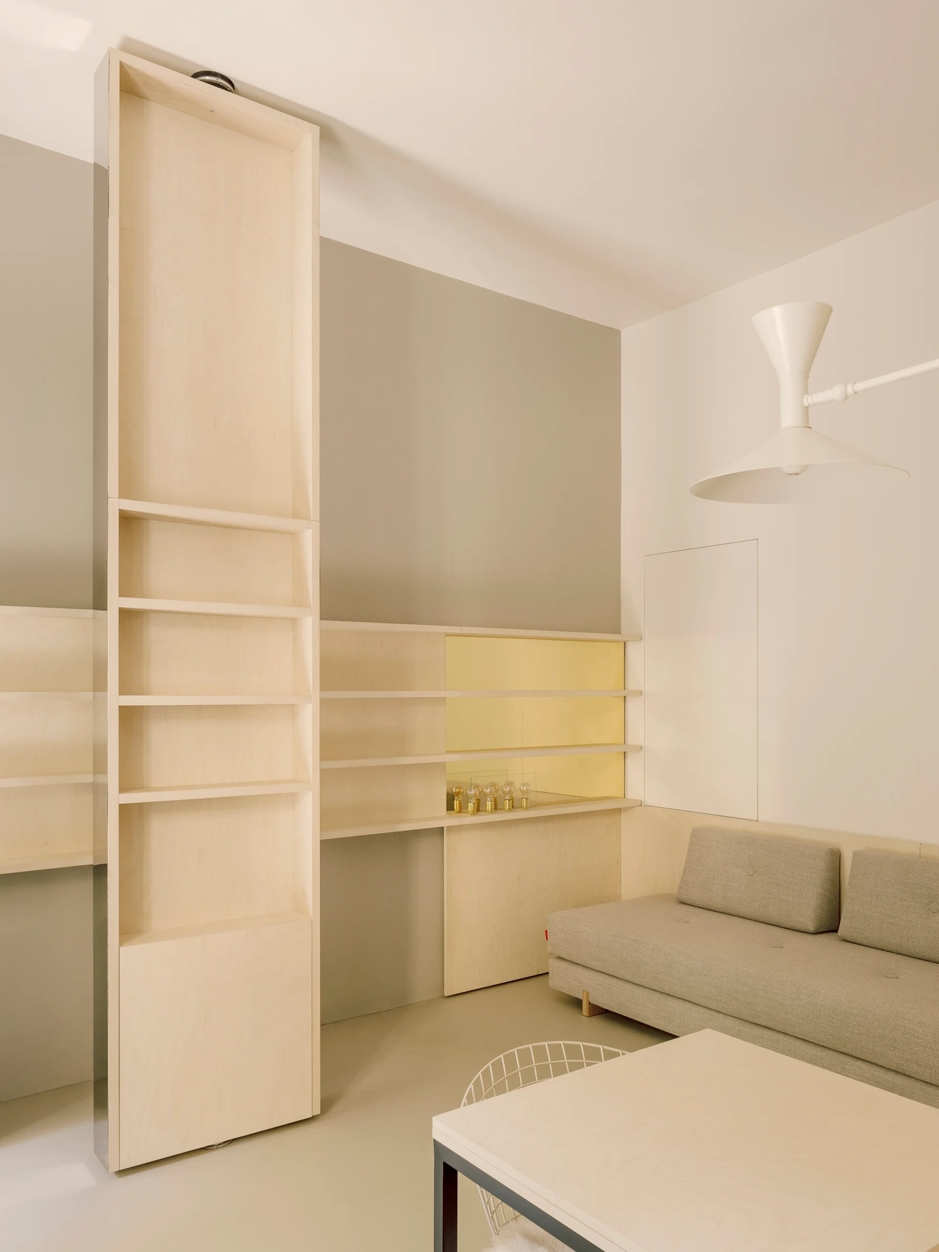 A minimalist living room features custom light wood shelving, a grey sofa, a matching low wooden table, and a gold mirror insert behind the shelves.