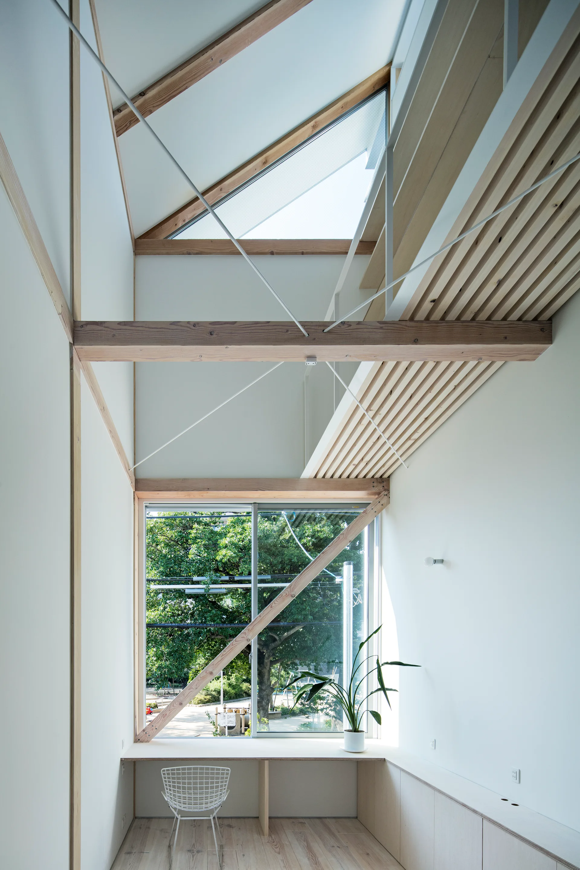 A minimalist workspace features a long built-in wooden desk under a large window overlooking trees, set beneath a vaulted ceiling with light wood beams.