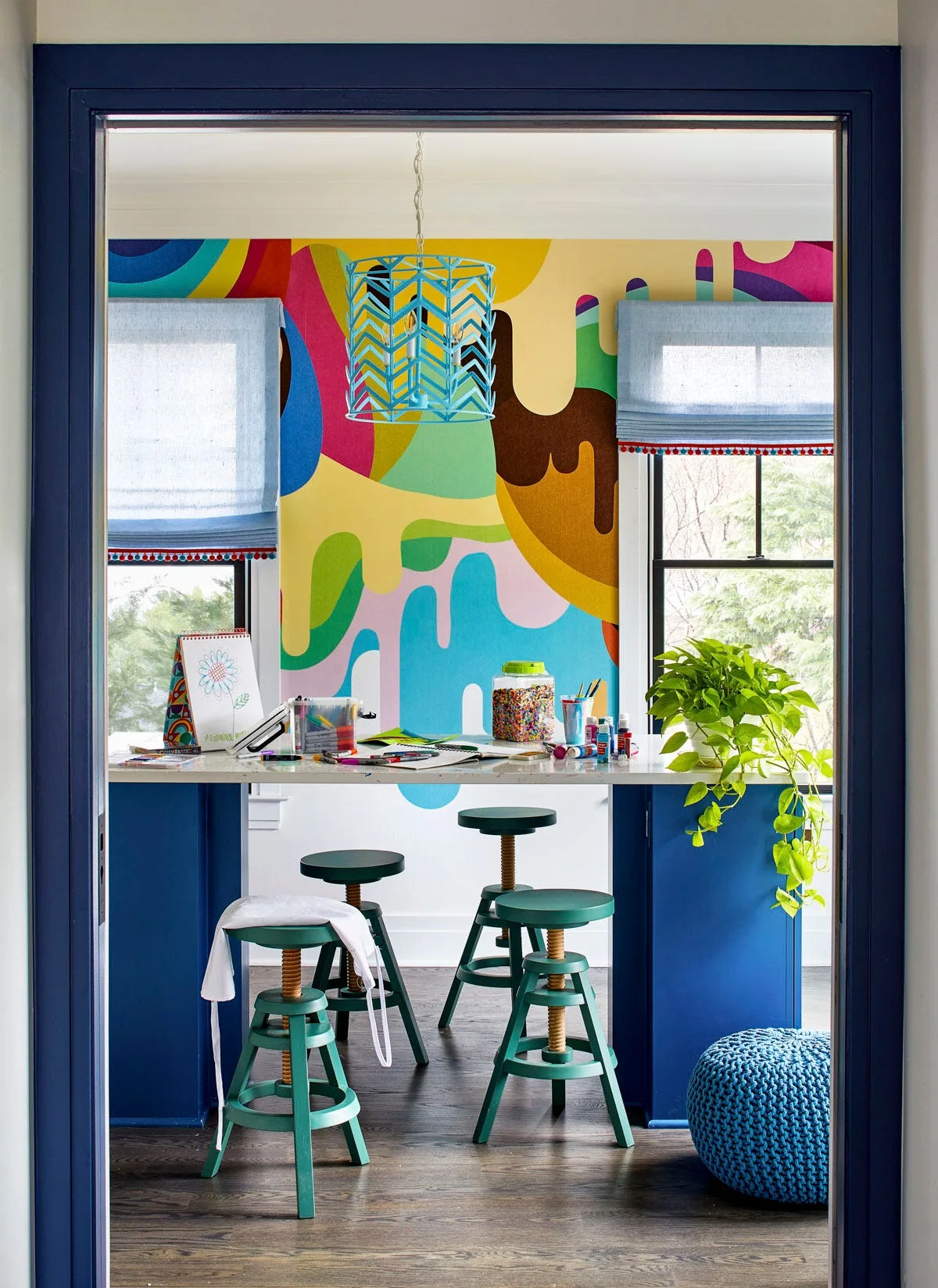 A kids' craft room viewed through a dark blue doorway features a colorful abstract wall mural, a white work table with green stools, and blue Roman shades.