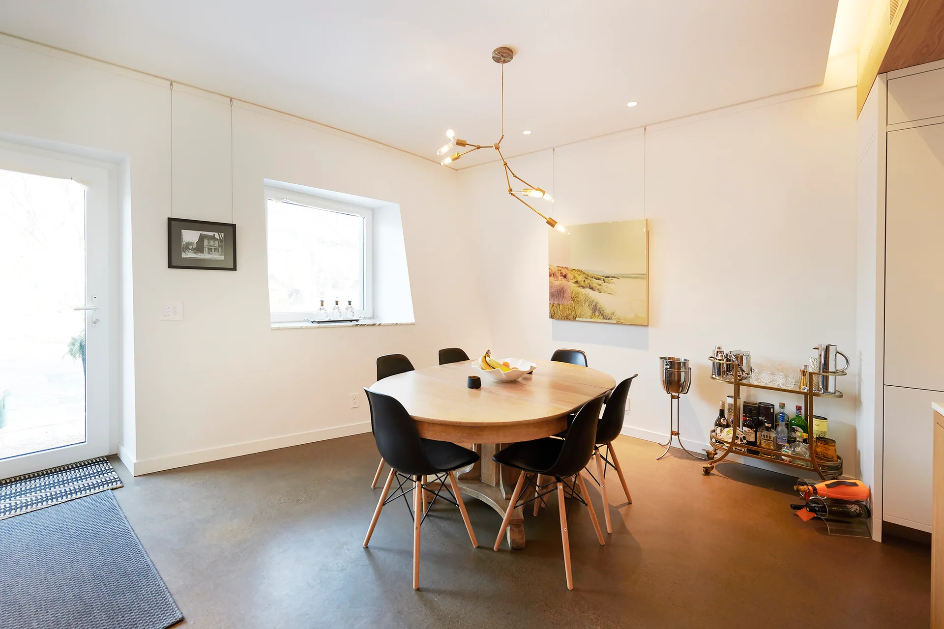 A round wooden dining table with black shell chairs sits under a branching gold pendant light in a room with white walls and concrete floors.