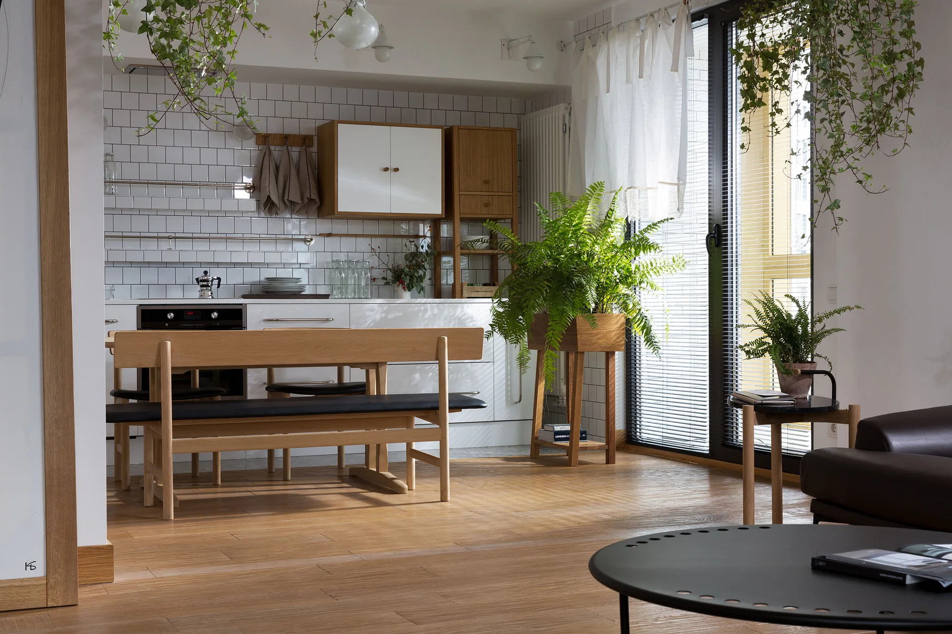 A modern open kitchen features a light wood dining bench, white subway tile backsplash, and abundant green houseplants near a large window.