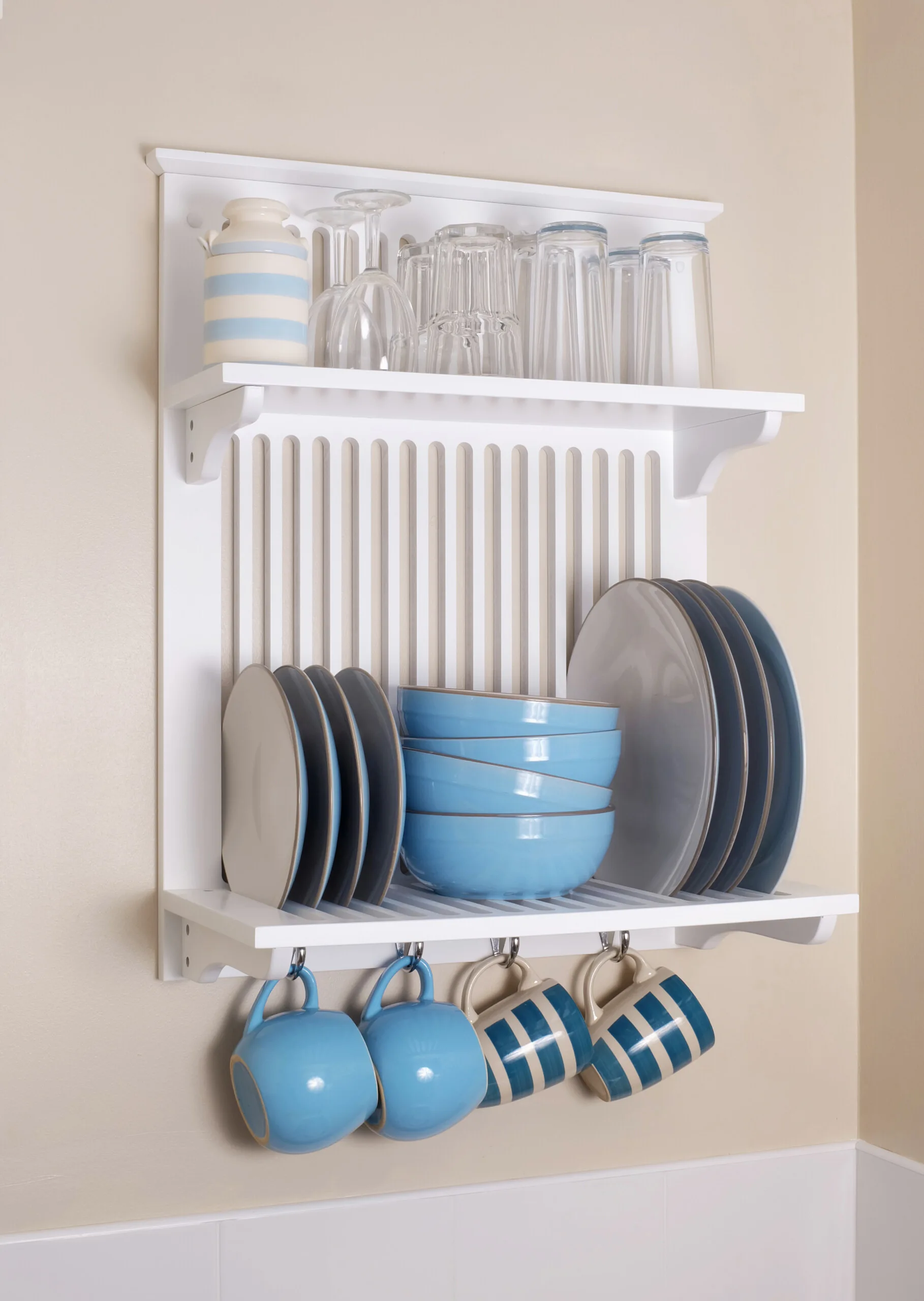 A white wooden wall-mounted plate rack holds blue bowls, grey plates, and glassware, with striped blue mugs hanging from hooks below.