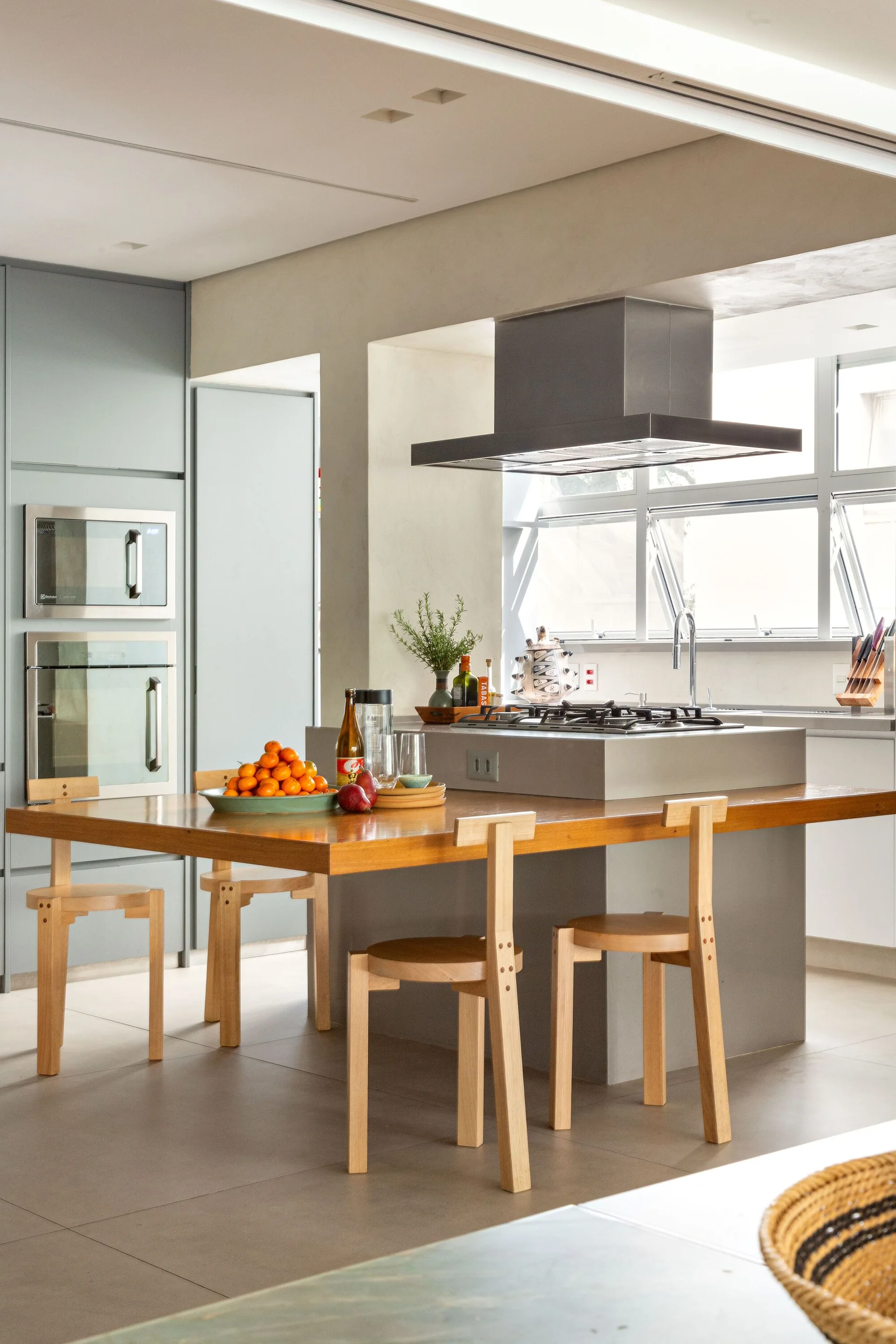 A modern grey kitchen island with an integrated cooktop and an attached warm wood dining table surrounded by light wood chairs.