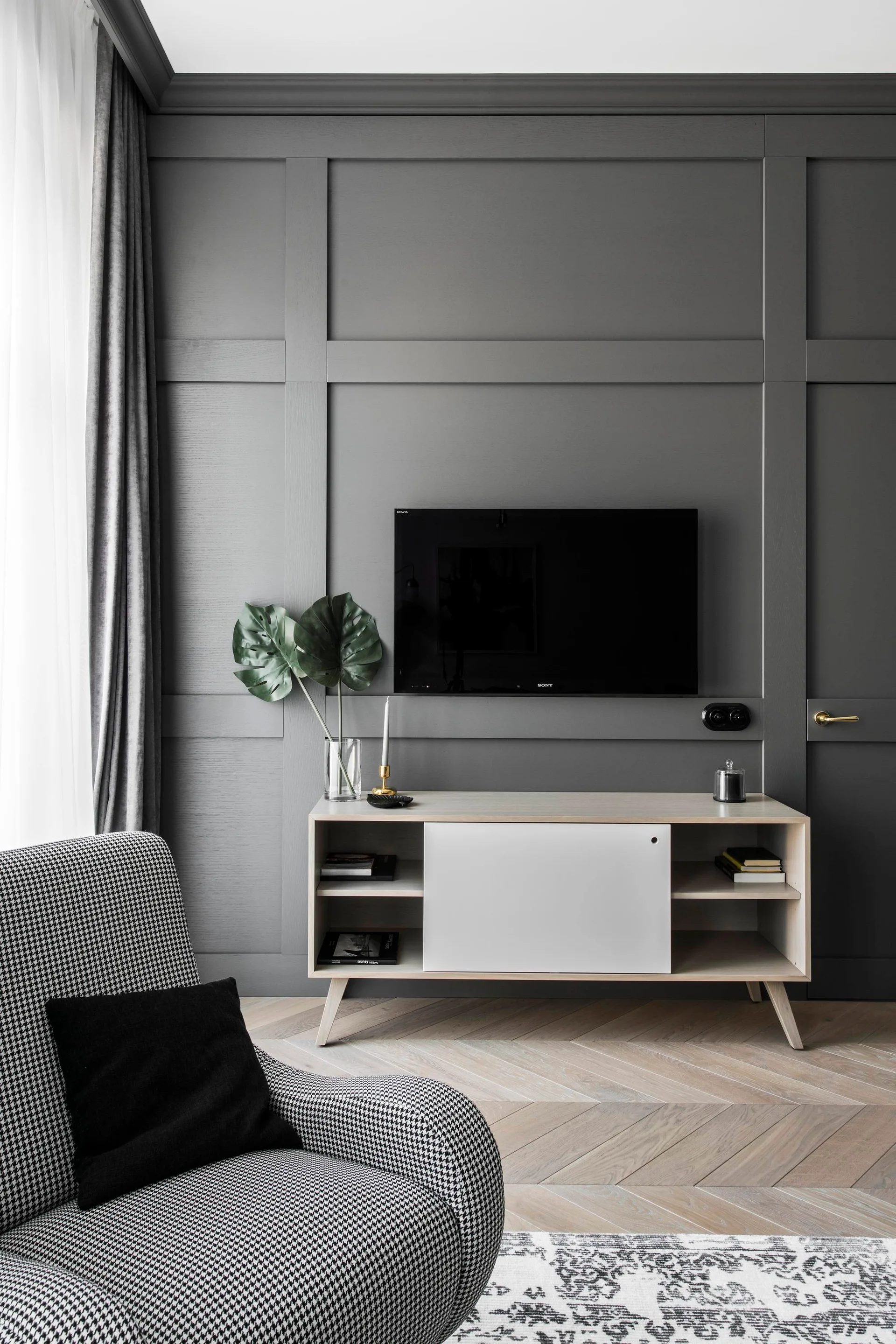 A living room corner features a dark grey paneled wall with a mounted television above a light wood media console and a houndstooth patterned armchair.