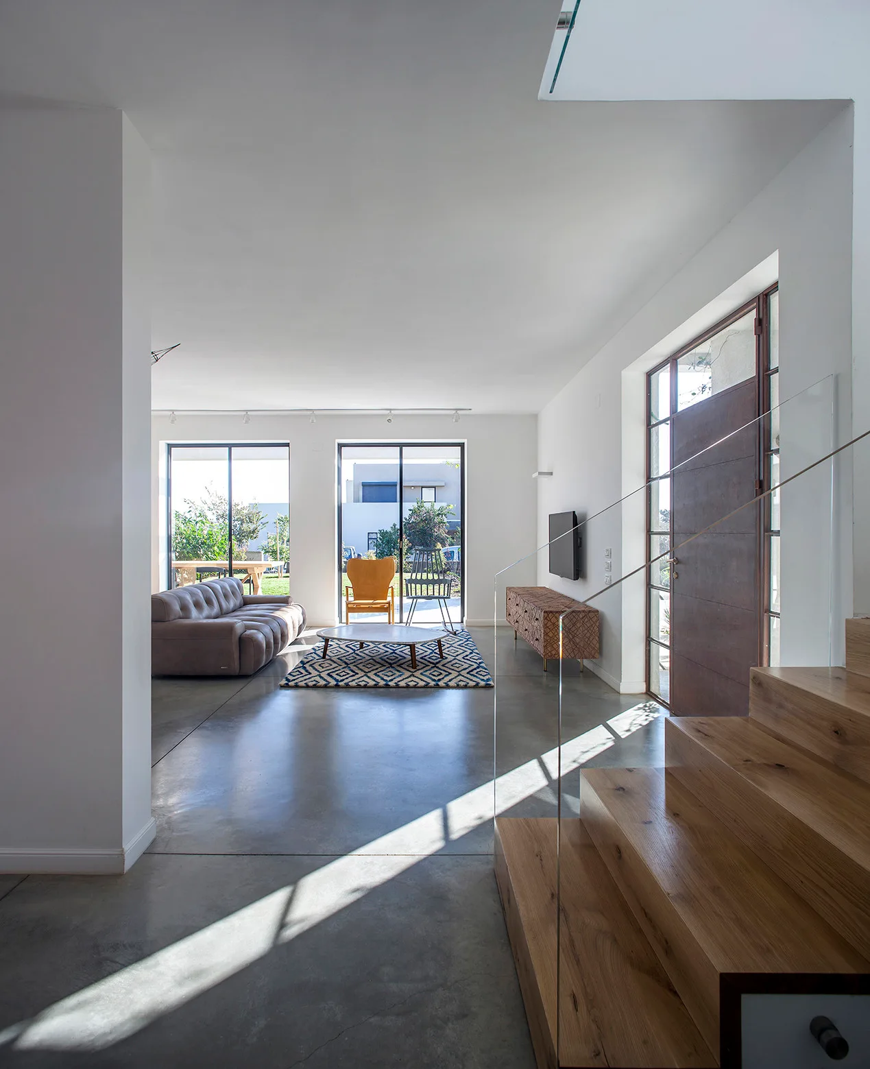 A modern living room features a wooden staircase with glass railing, polished concrete floors, a grey leather sofa, and large glass patio doors.