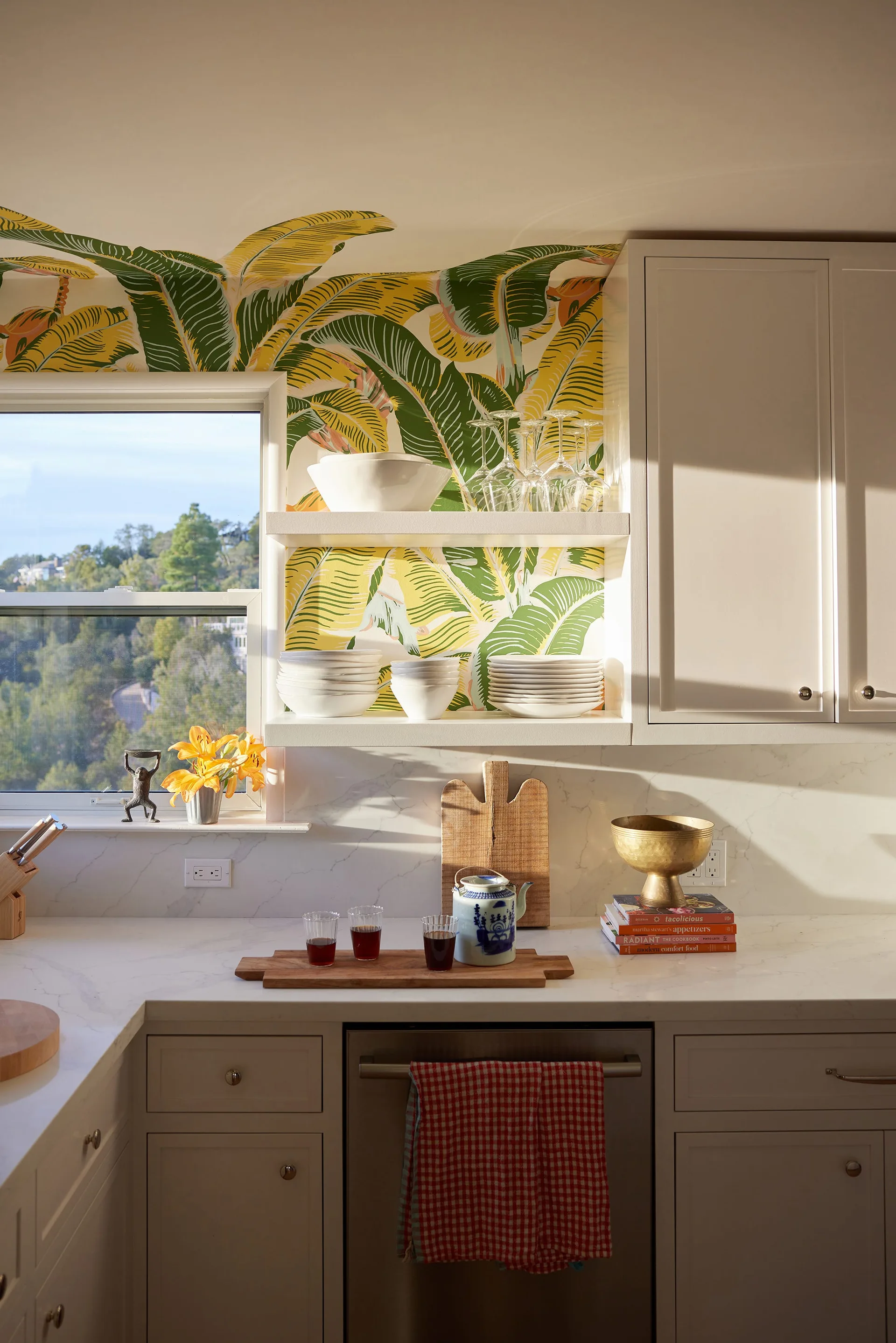 White kitchen cabinets and open shelves displaying dishware against green and yellow tropical leaf wallpaper next to a window.