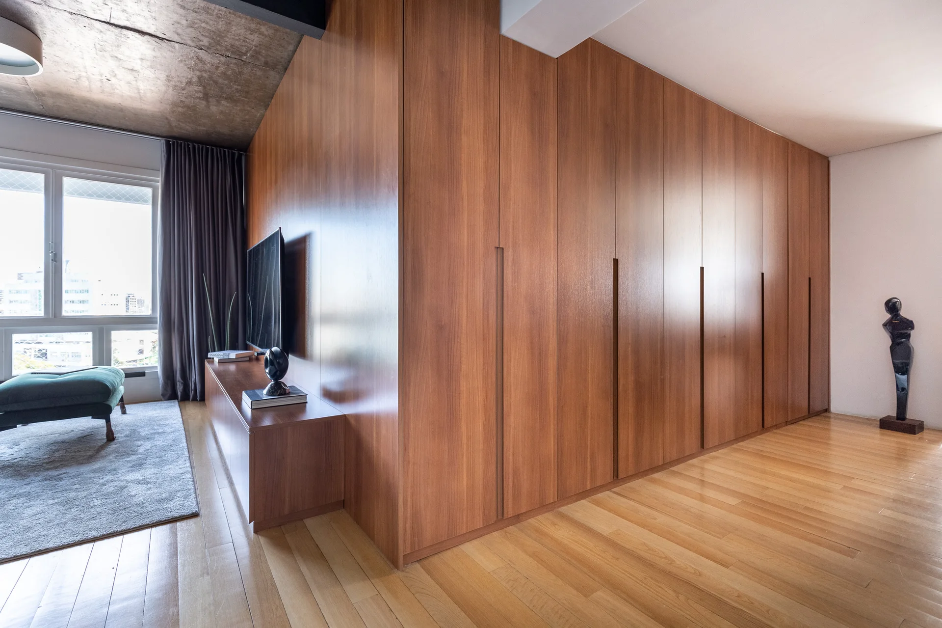 A modern living room features a floor-to-ceiling wood storage cabinet system next to a low media console, hardwood floors, and an exposed concrete ceiling.
