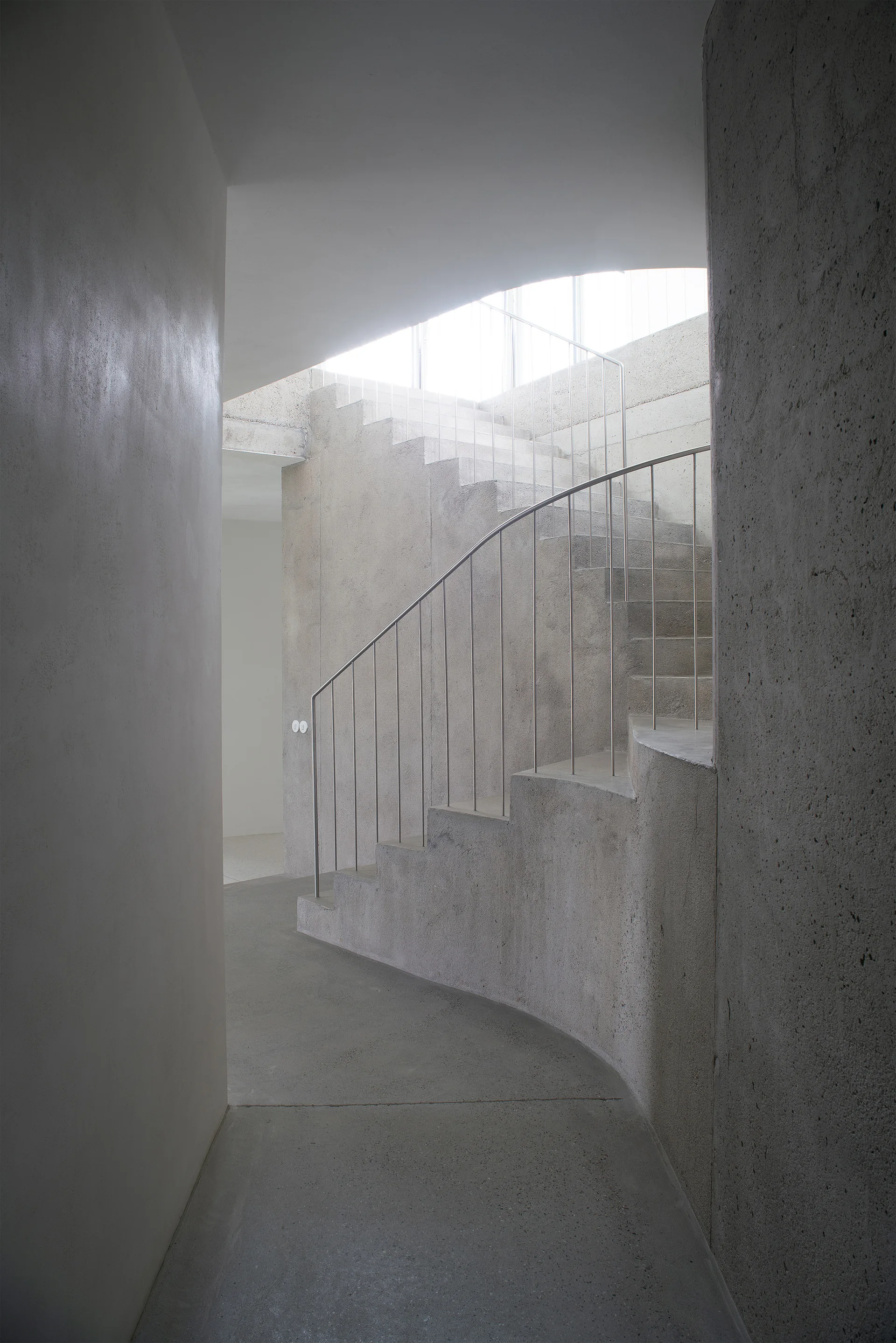 A curved concrete staircase with a slender metal handrail rises through a monochromatic grey hallway illuminated by soft overhead light.