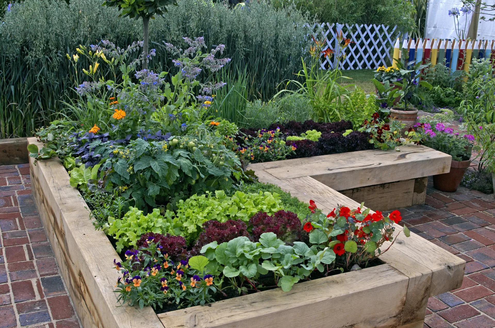 An L-shaped raised garden bed made of thick wooden timber beams sits on a red brick patio filled with green vegetables and flowers.