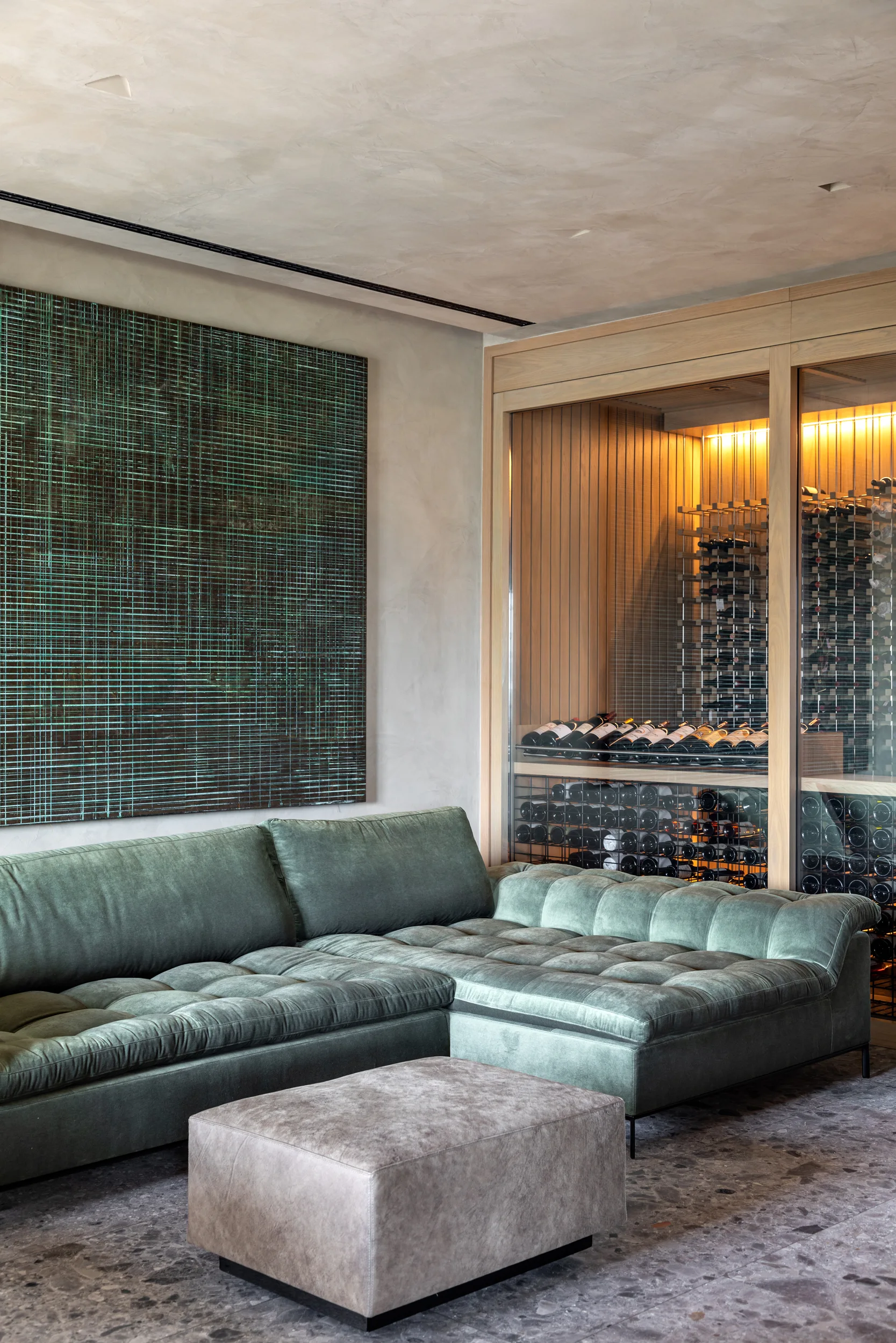 A contemporary living space featuring a tufted sage green velvet sectional, a square stone ottoman, and an illuminated glass-enclosed wine cellar.