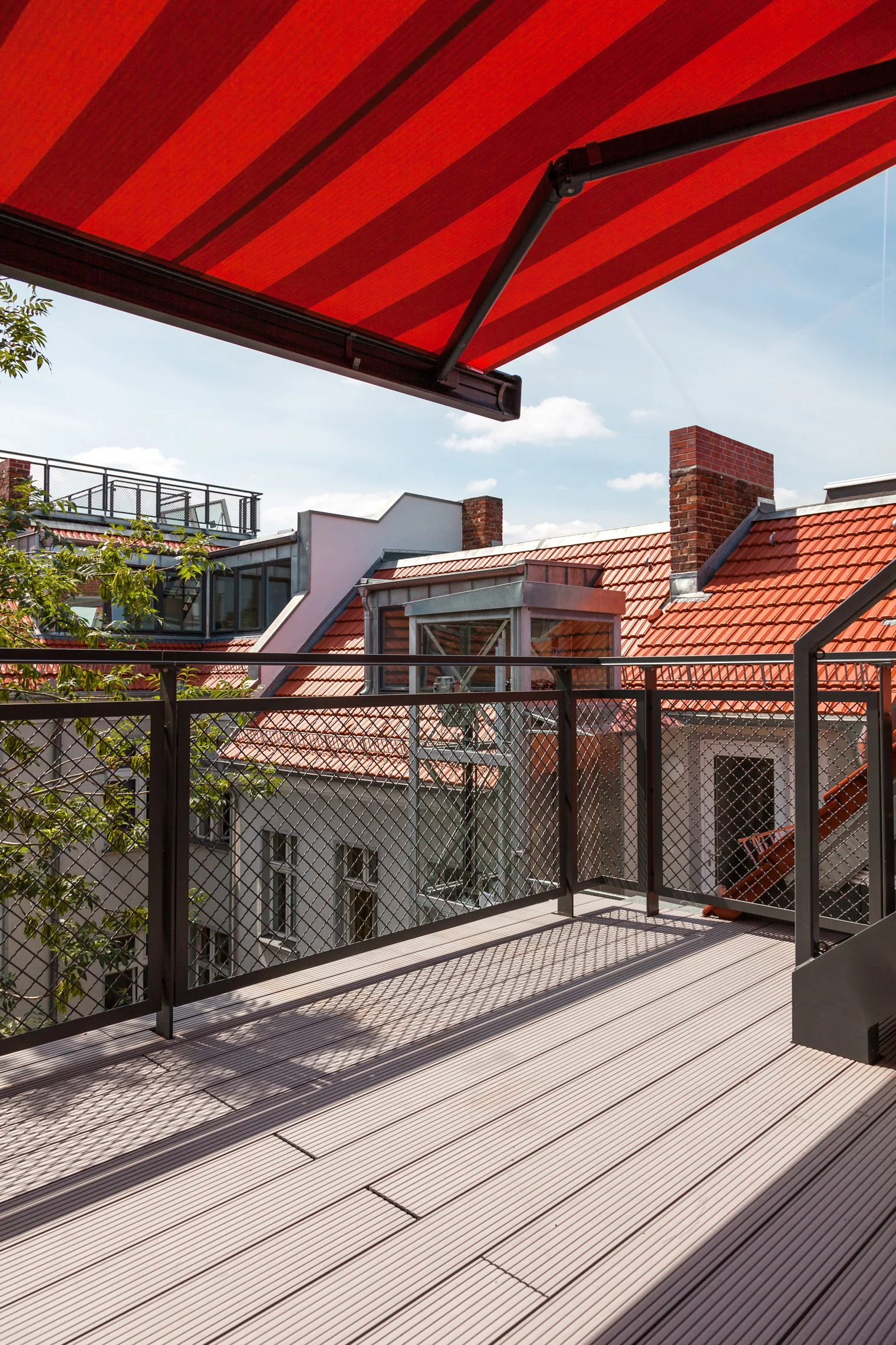 An outdoor rooftop terrace features light brown composite decking, a dark metal mesh railing, and a retractable red striped awning overhead.