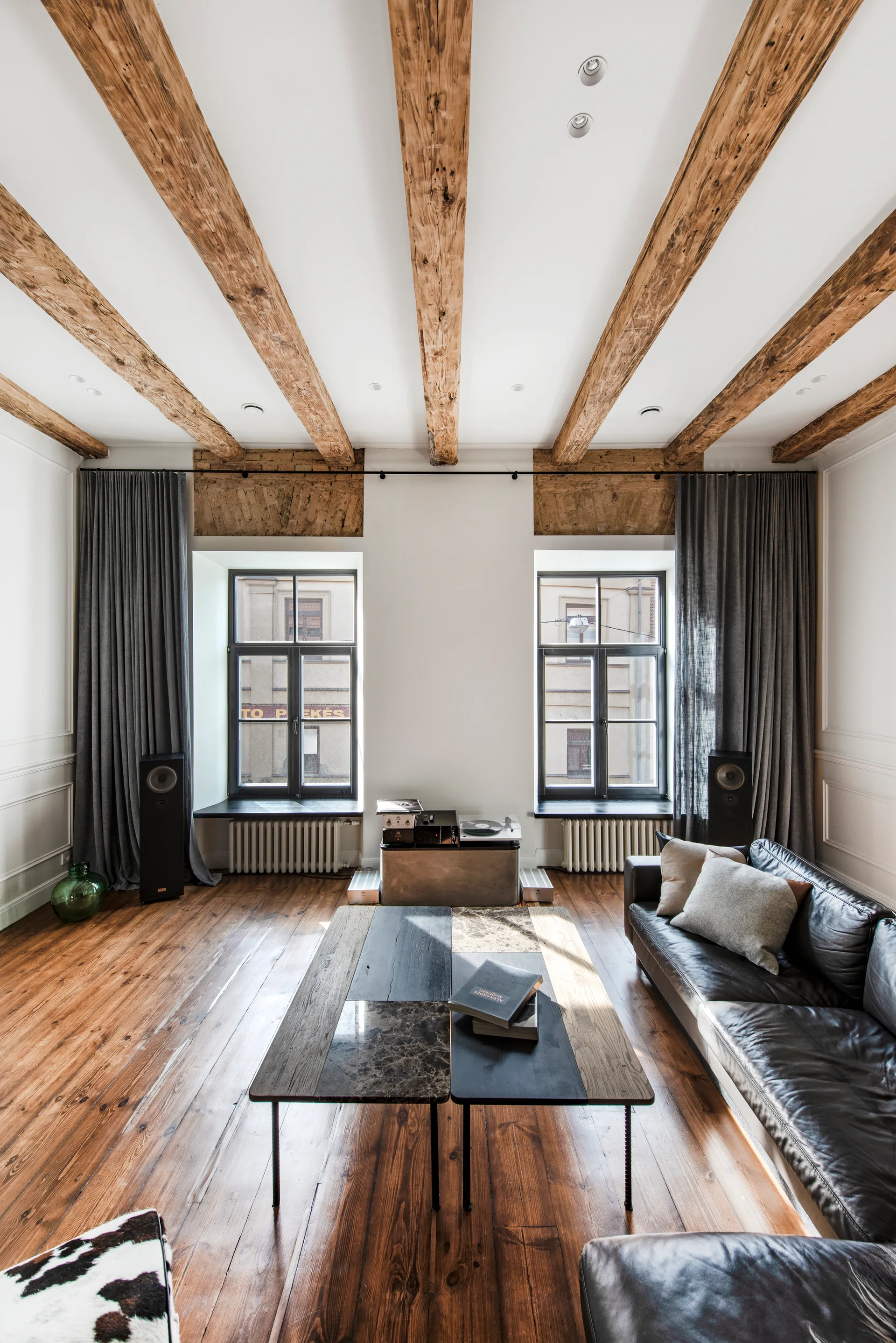 A living room features exposed wooden ceiling beams, a black leather sofa, a multi-material coffee table, and tall windows framed by grey curtains.