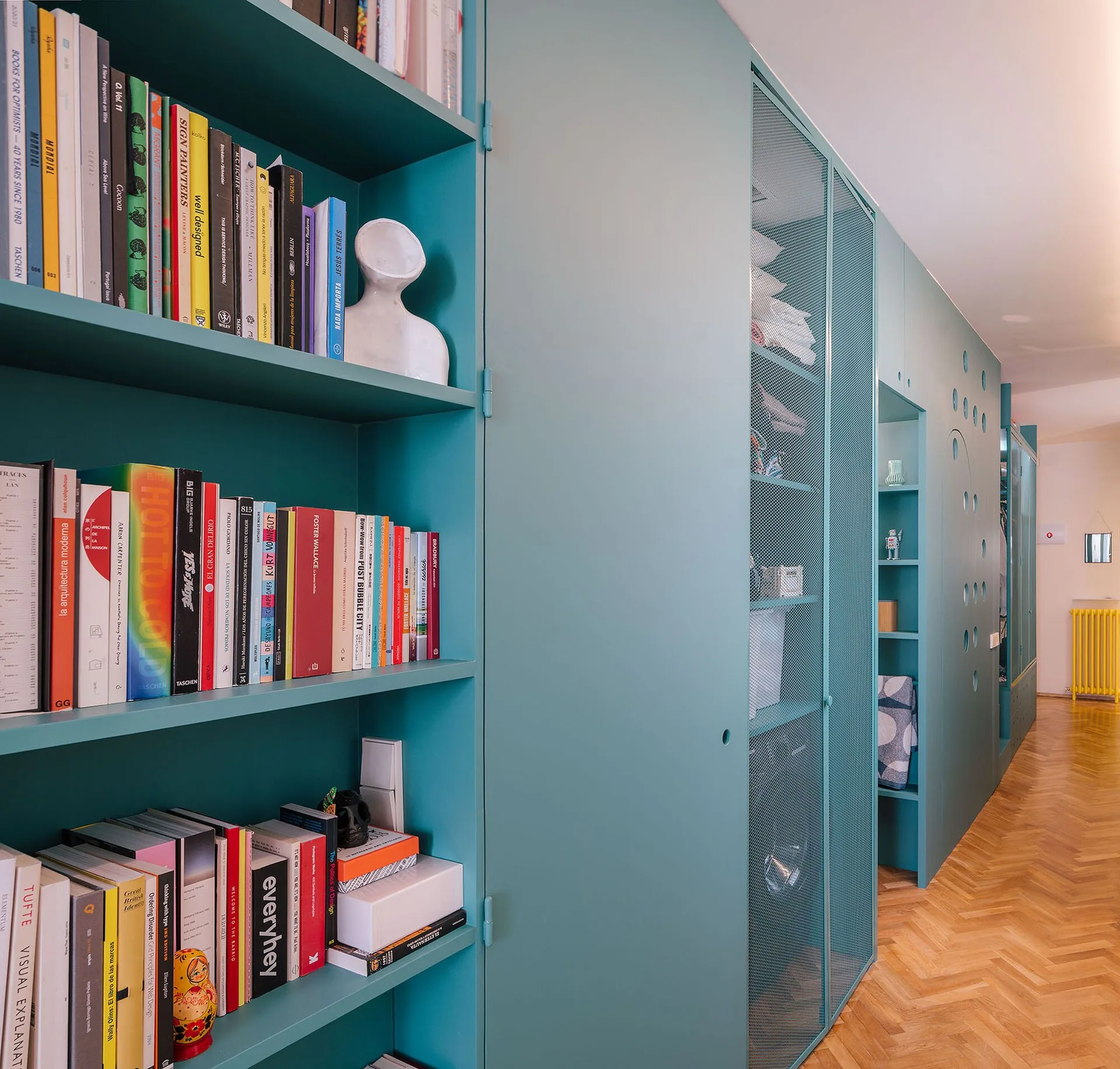 Floor-to-ceiling teal cabinetry lines a hallway with a herringbone wood floor, featuring built-in bookshelves and integrated metal mesh doors.