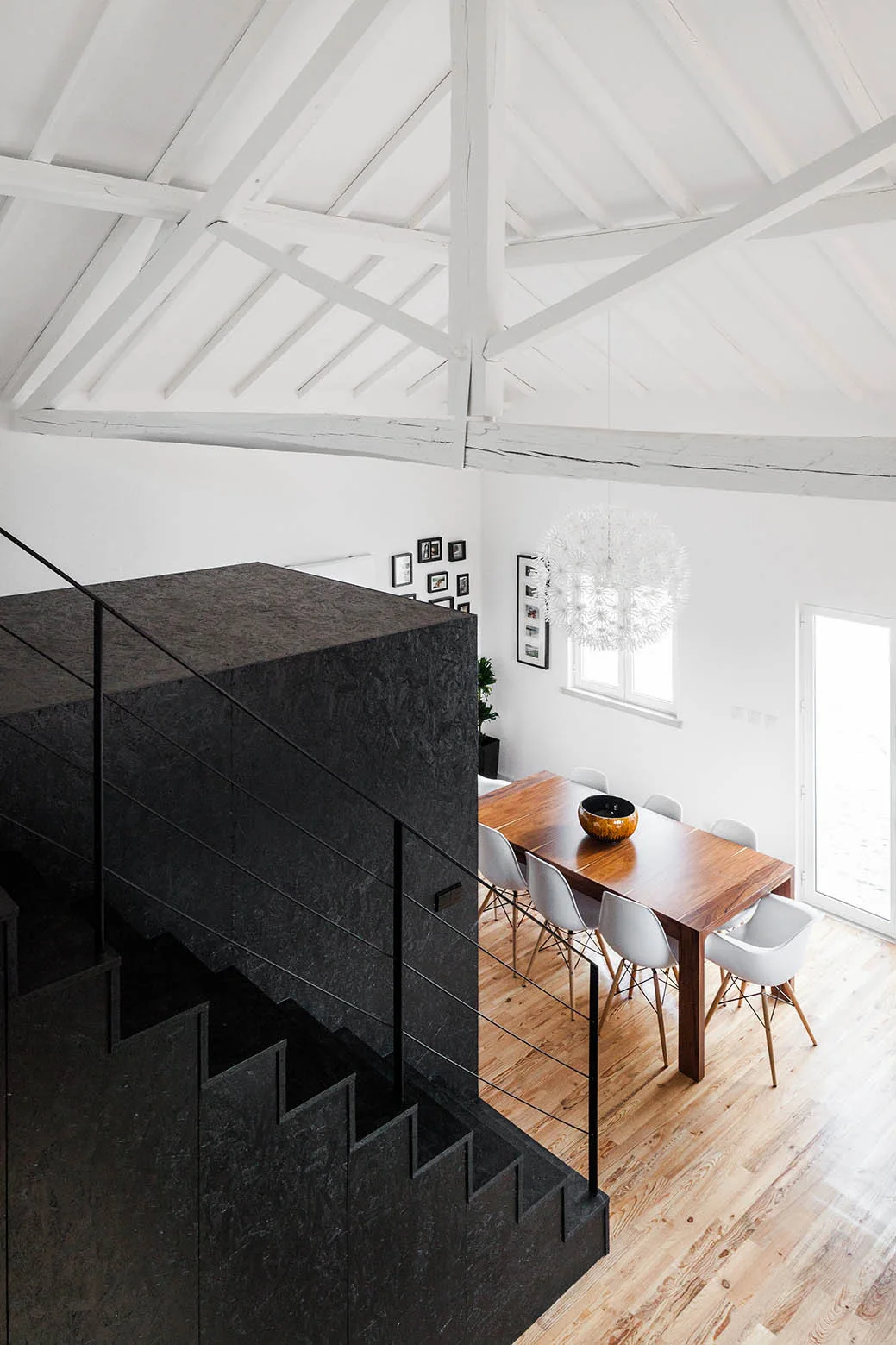A high-angle view of a loft dining room featuring a wood table, white chairs, a black textured staircase enclosure, and white vaulted ceiling beams.