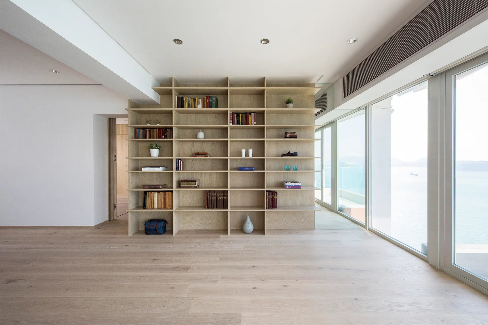 A minimalist room featuring a large, floor-to-ceiling light wood bookshelf next to glass doors showing a coastal water view.