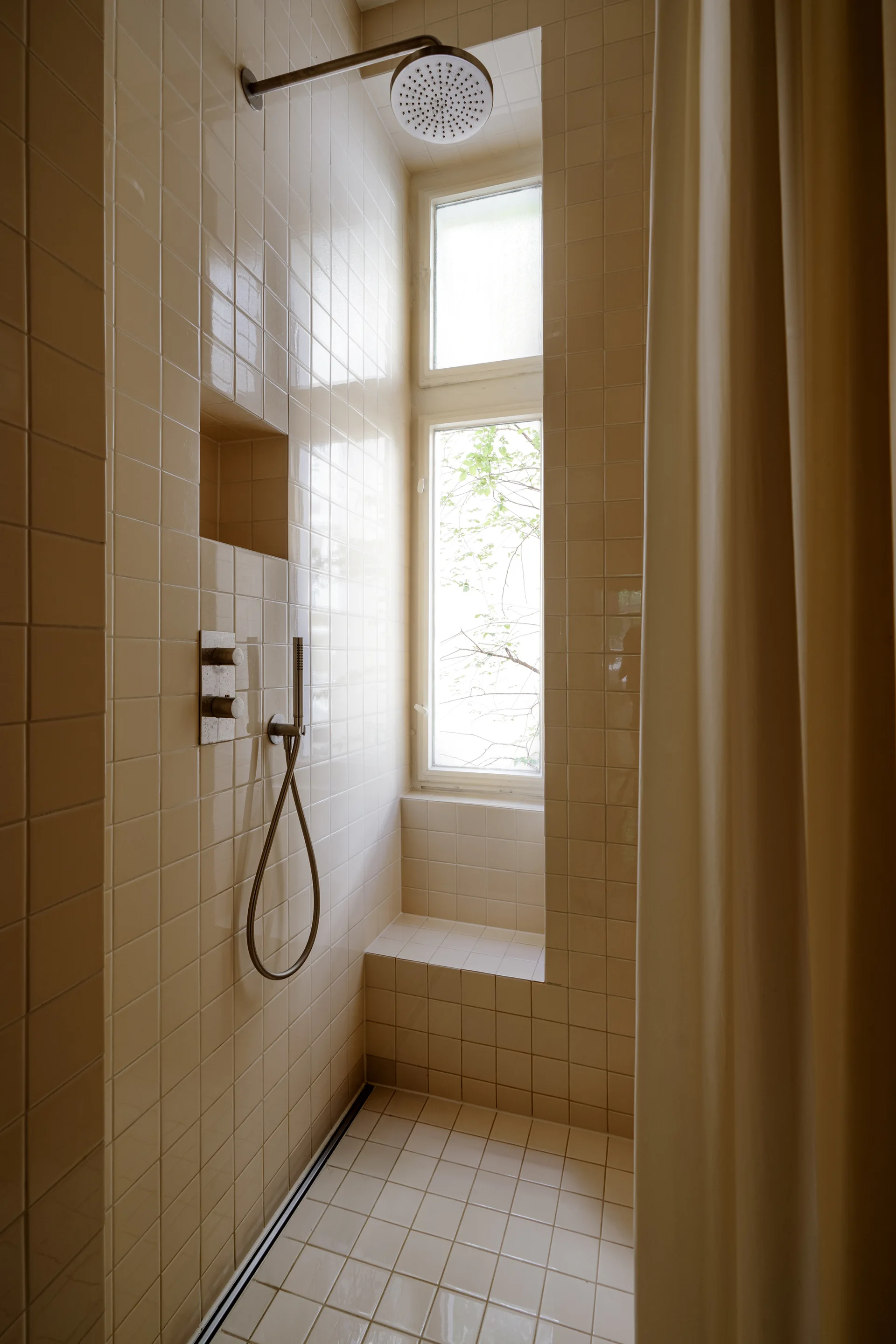 A compact shower stall features glossy beige square tiles on the walls and floor, a built-in tiled bench, and a tall window looking outside.