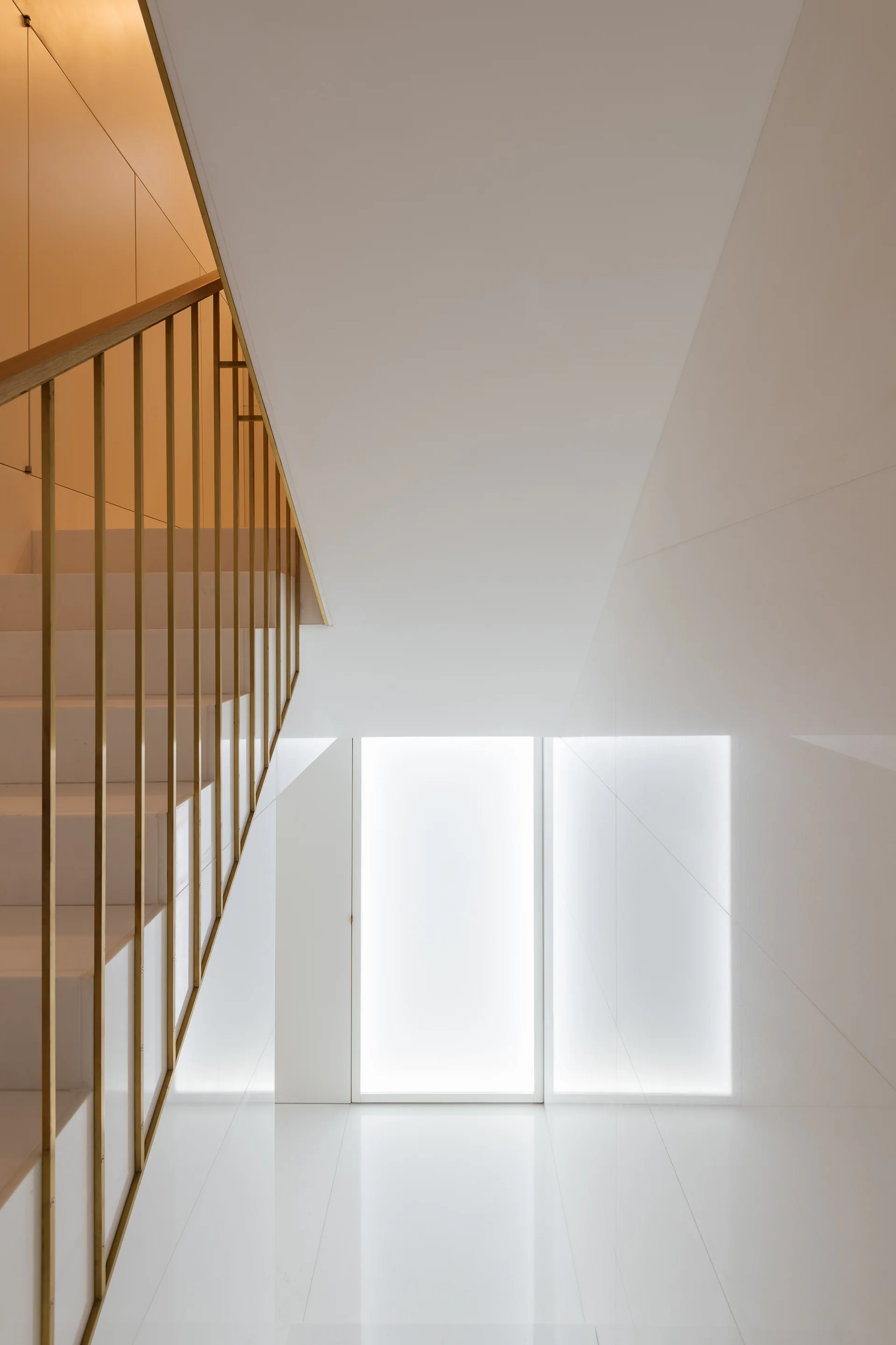 A minimalist white staircase features a slim gold metal railing and leads down to a glossy floor lit by a backlit glass panel.