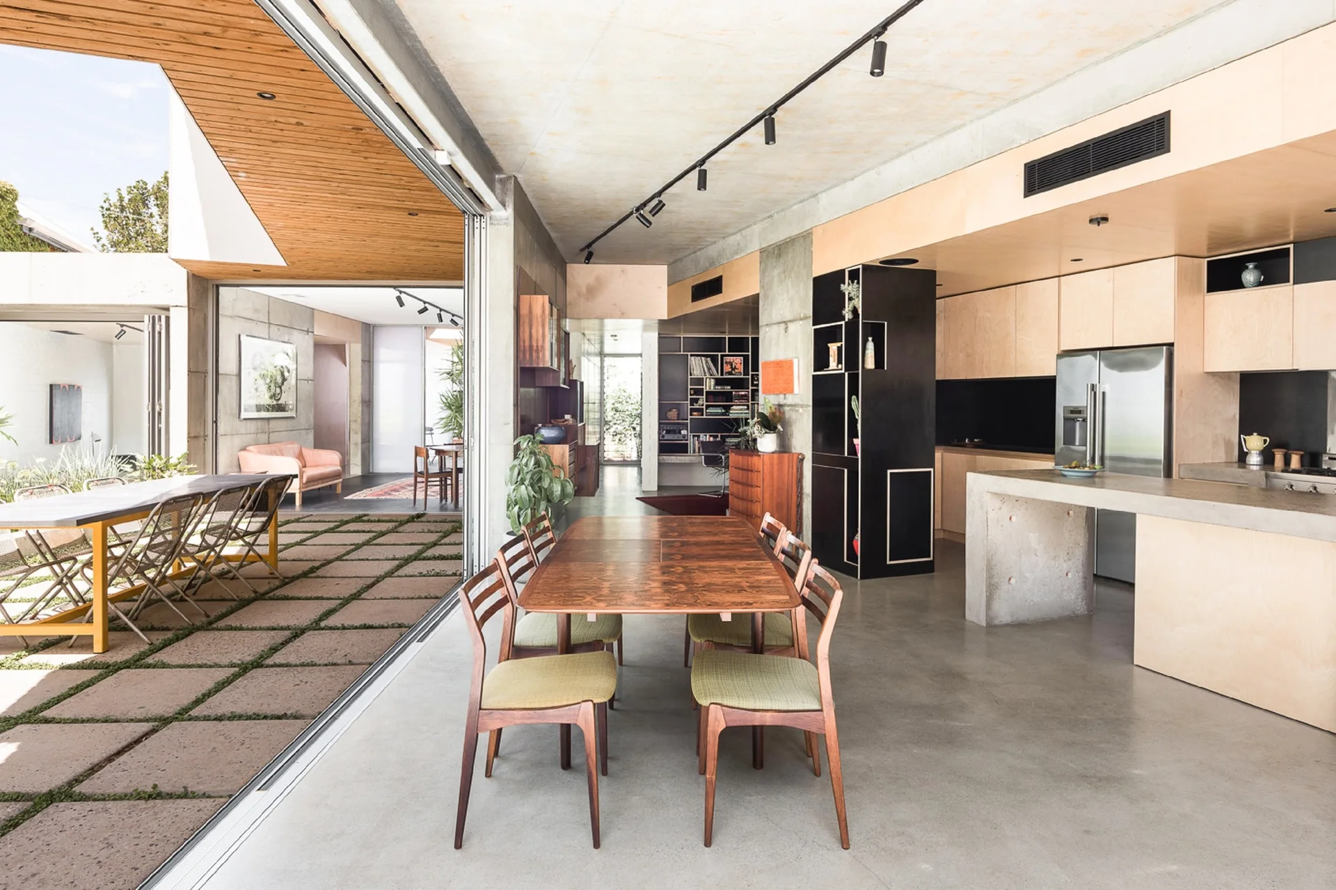 A modern open-concept dining room and kitchen feature raw concrete walls, warm wood cabinetry, and sliding doors opening to an outdoor terrace.
