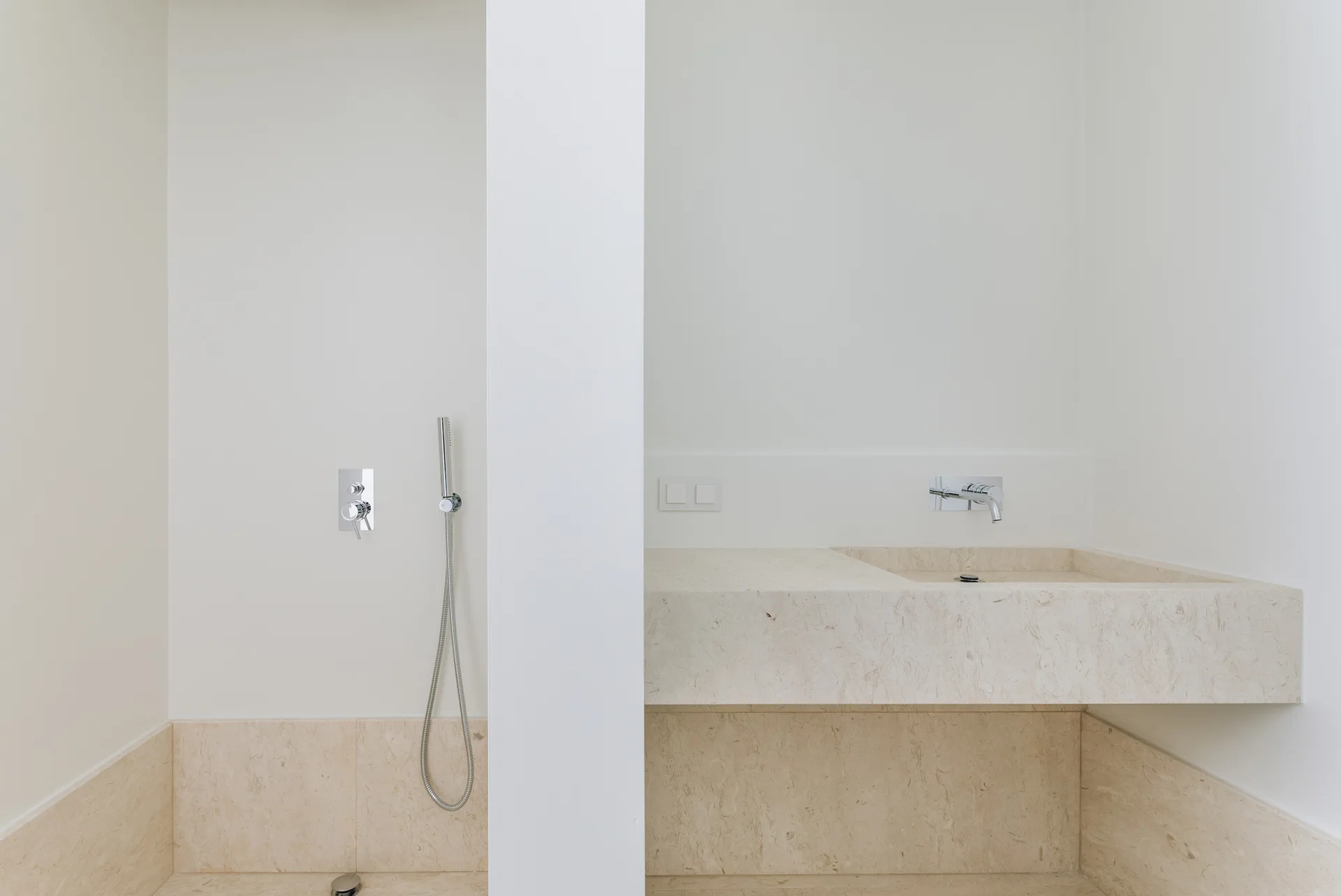 A minimalist bathroom features a floating beige stone vanity with an integrated sink next to a white partition wall and chrome shower fixtures.