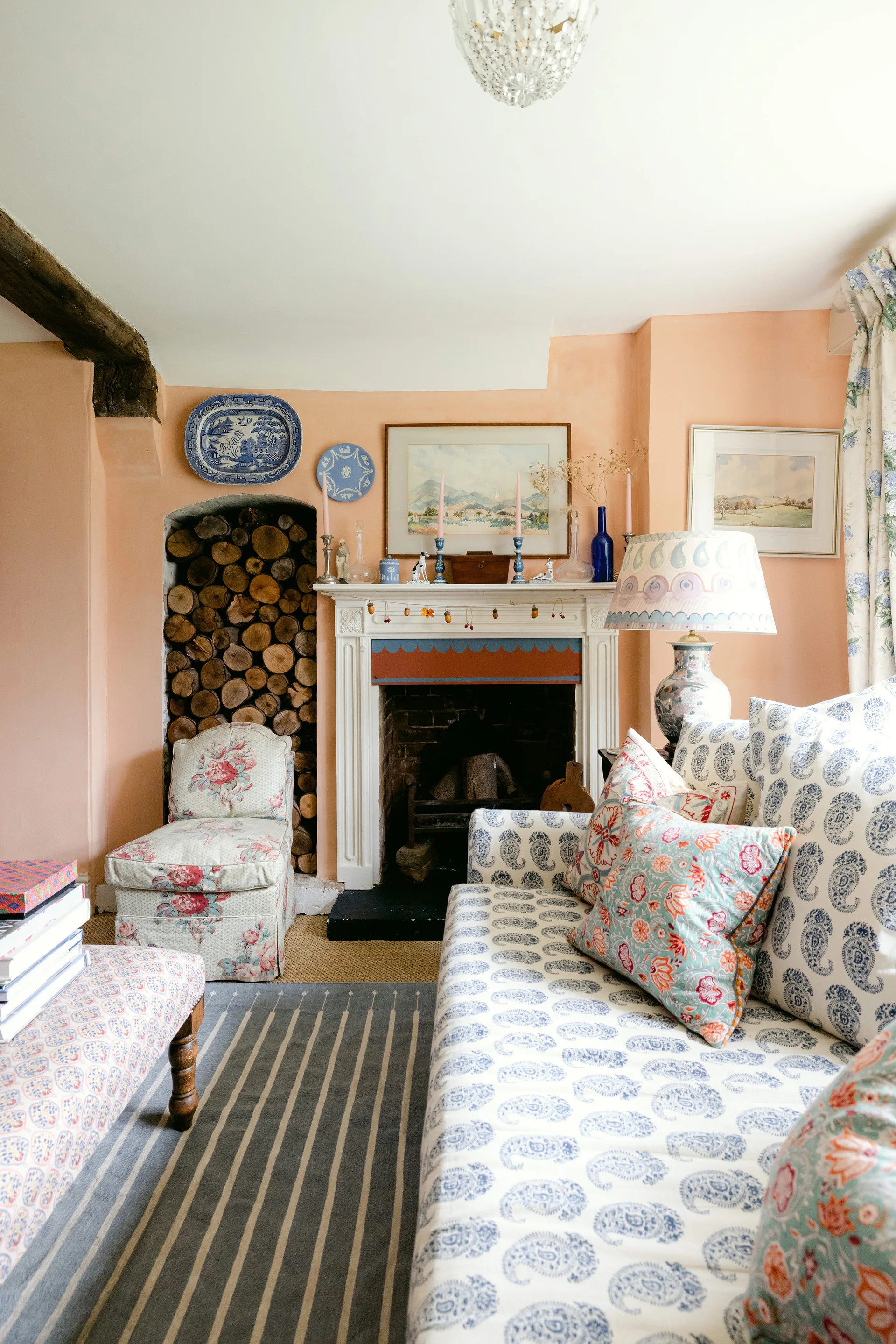A cozy living room featuring peach walls, a white fireplace next to an alcove filled with firewood, and a sofa upholstered in blue paisley fabric.