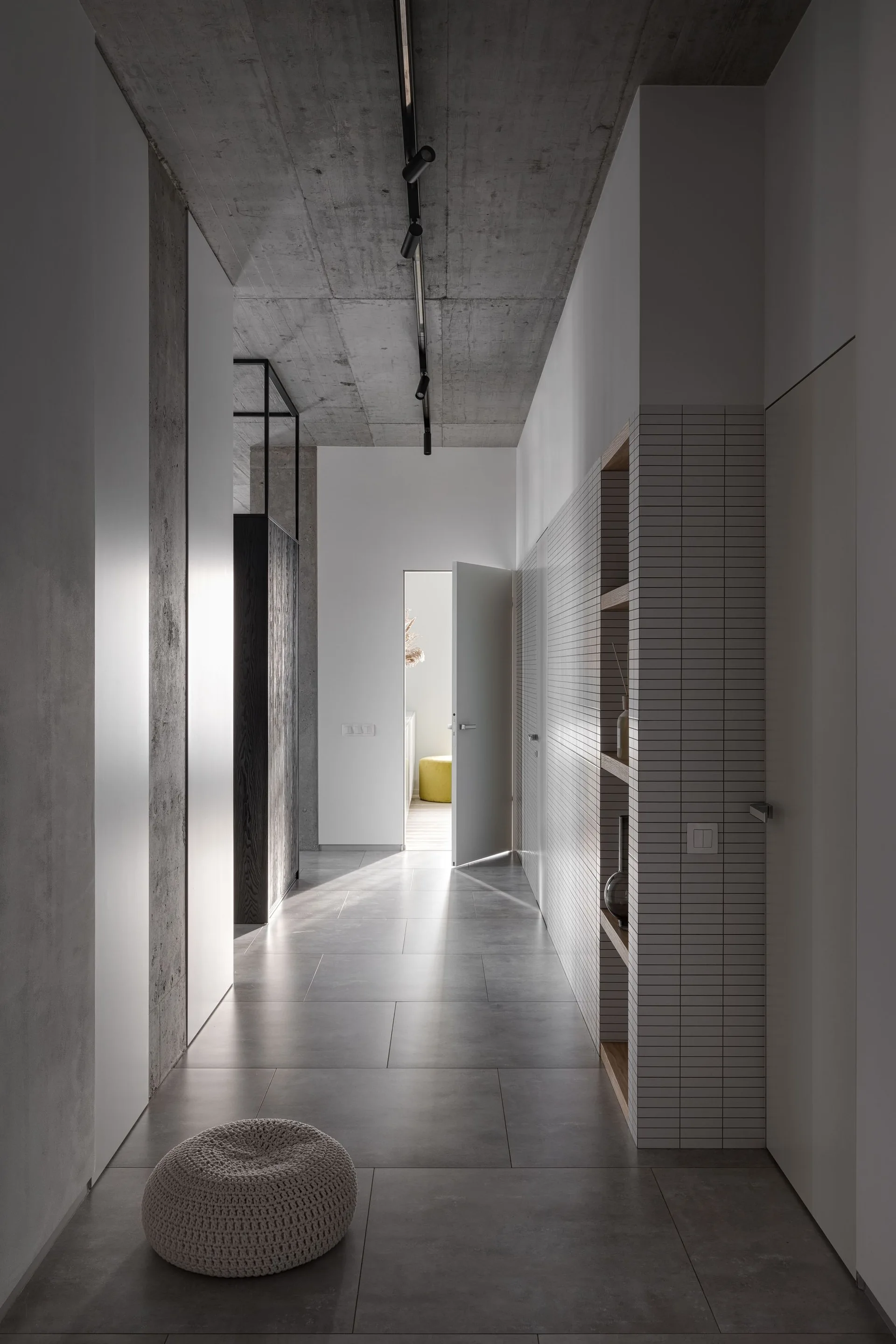 A minimalist hallway featuring an exposed concrete ceiling, a white grid-tiled wall with recessed wooden shelves, and large grey tile floors.
