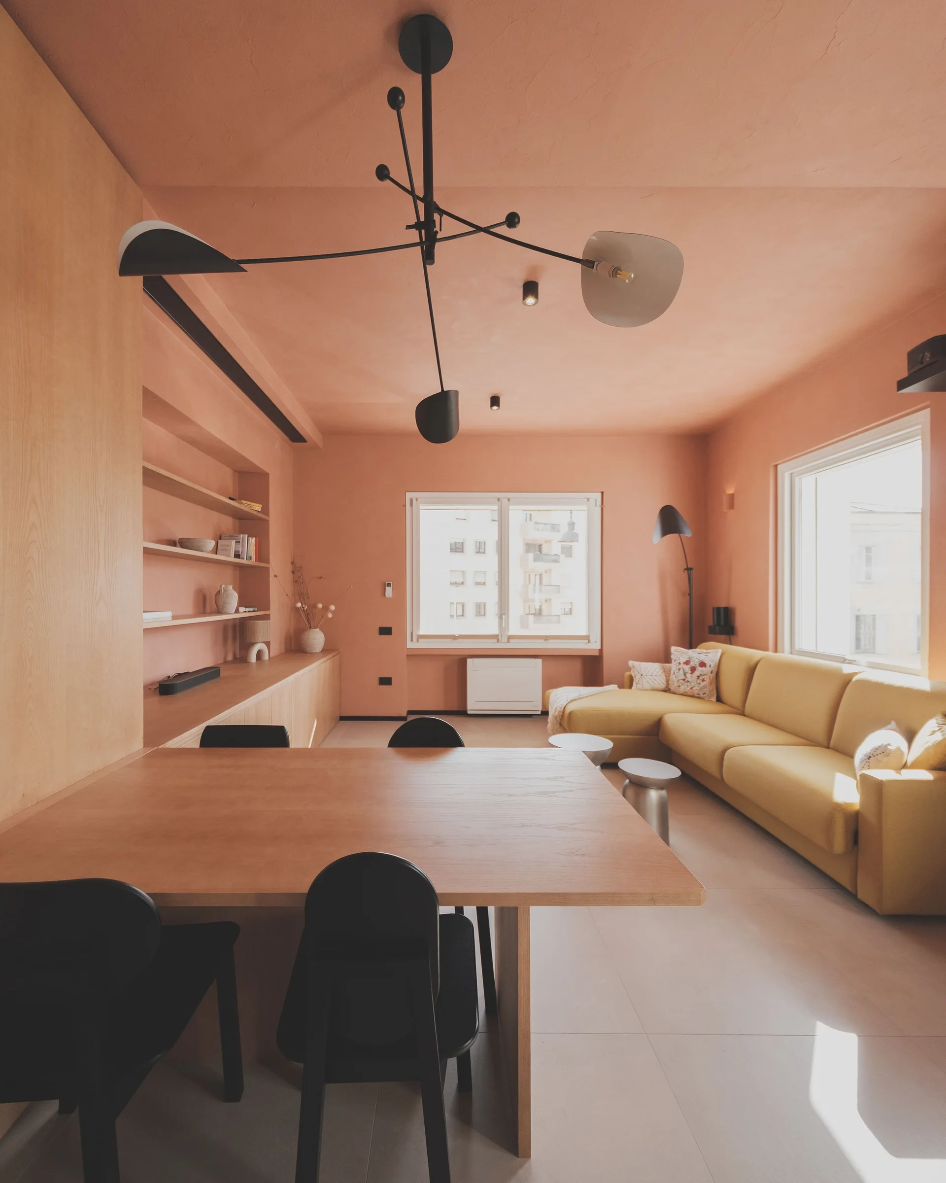 A contemporary living and dining area with peach walls, a yellow sectional sofa, a light wood dining table with black chairs, and built-in shelves.
