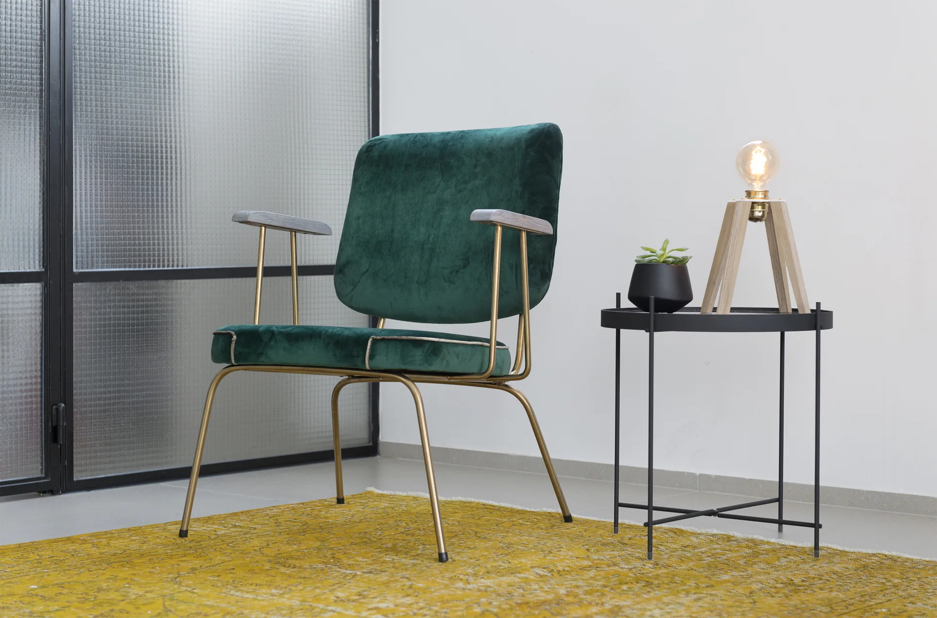 A green velvet armchair with a gold frame sits on a distressed yellow rug next to a black metal side table holding a modern lamp.
