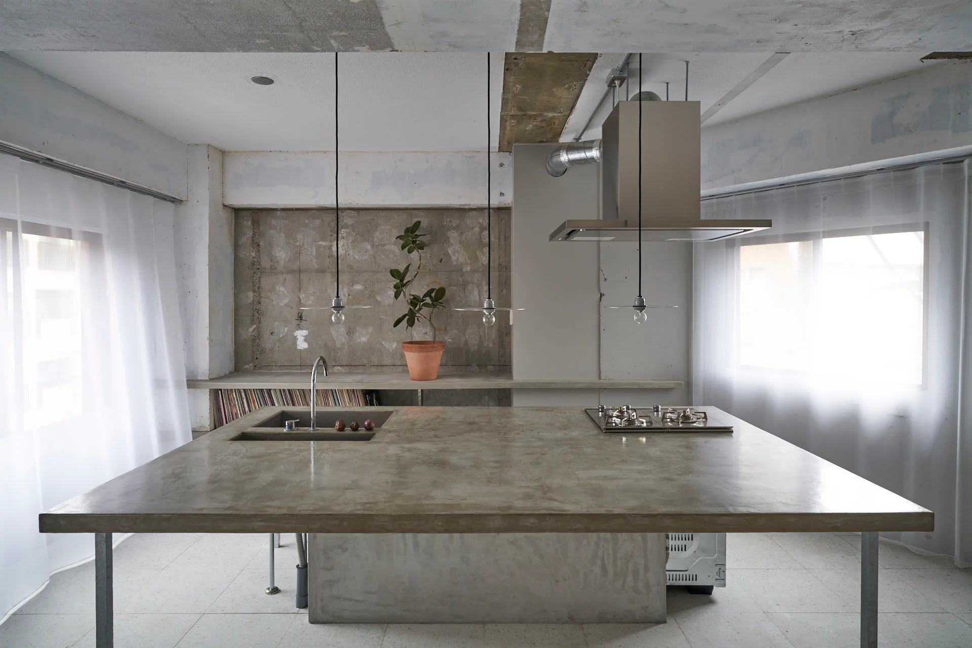 A minimalist industrial kitchen features a large raw concrete island with an integrated sink and cooktop beneath three simple pendant lights.
