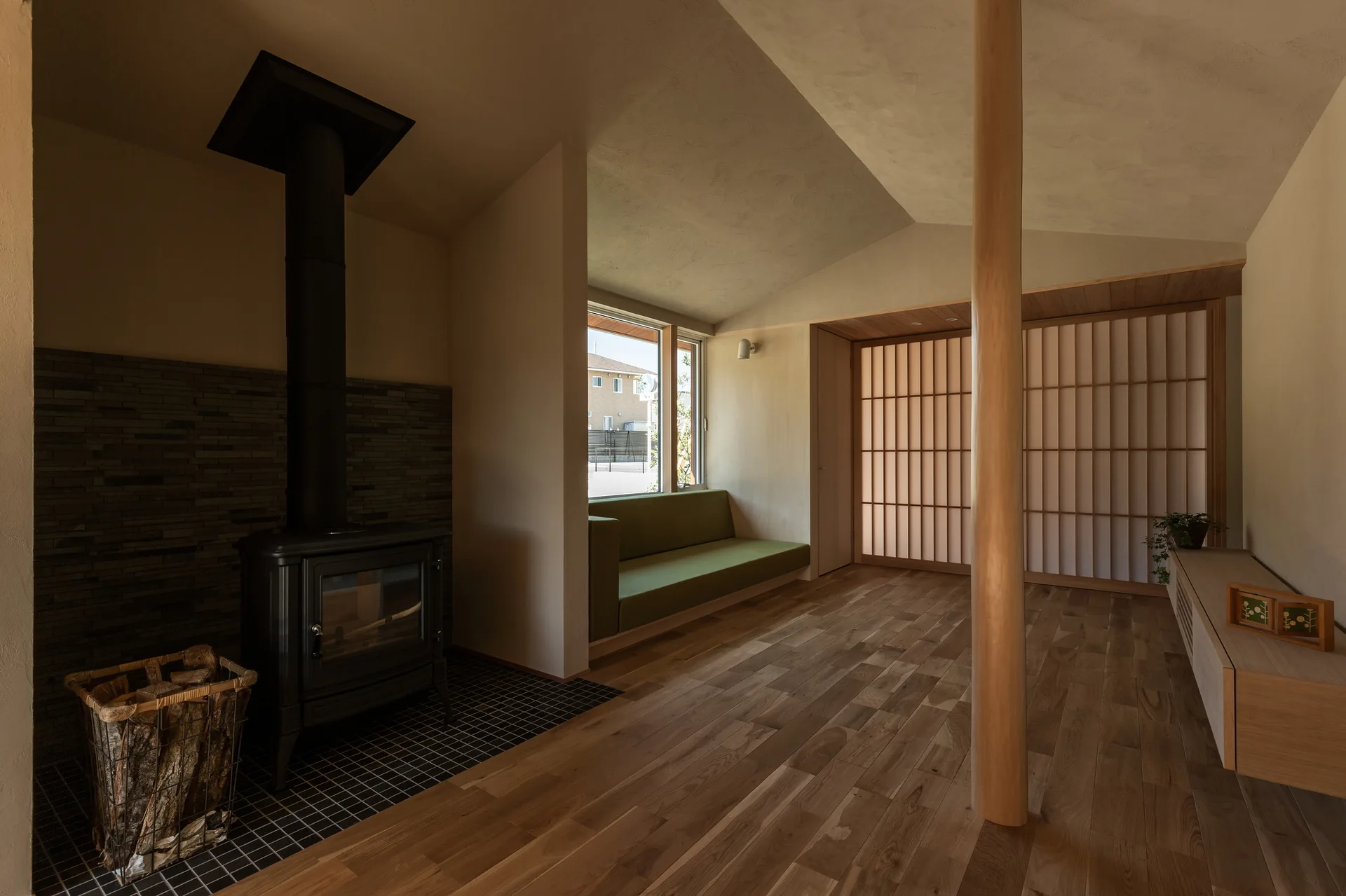 A minimalist living room features a black wood-burning stove, a built-in green bench sofa, a large wooden support pillar, and a shoji screen.