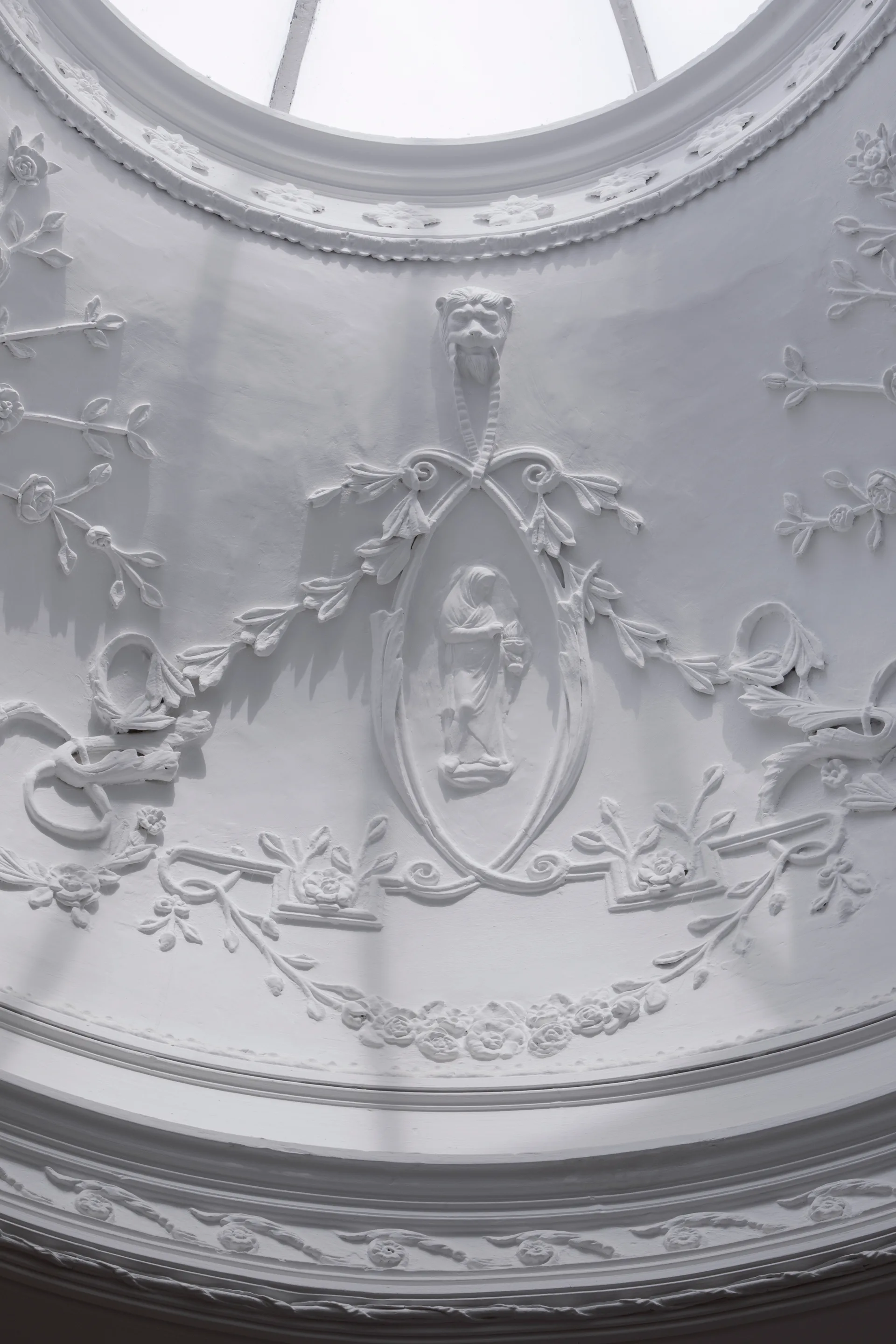 Detailed view of an ornate white plaster ceiling featuring intricate floral relief carvings, a central medallion, and a circular skylight above.
