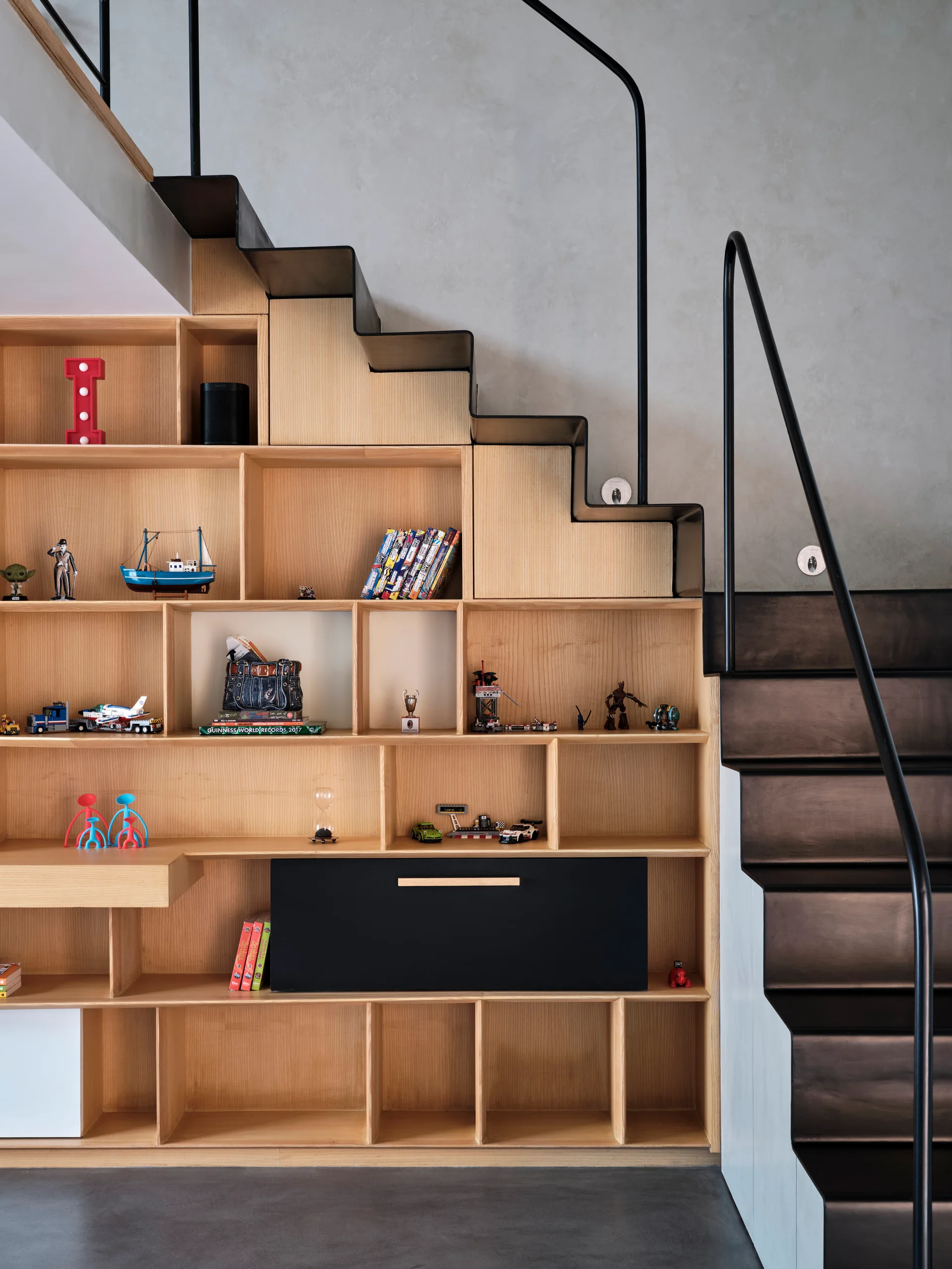A light wood built-in shelving unit filled with books and toys is integrated under a modern black metal staircase with a slim handrail.