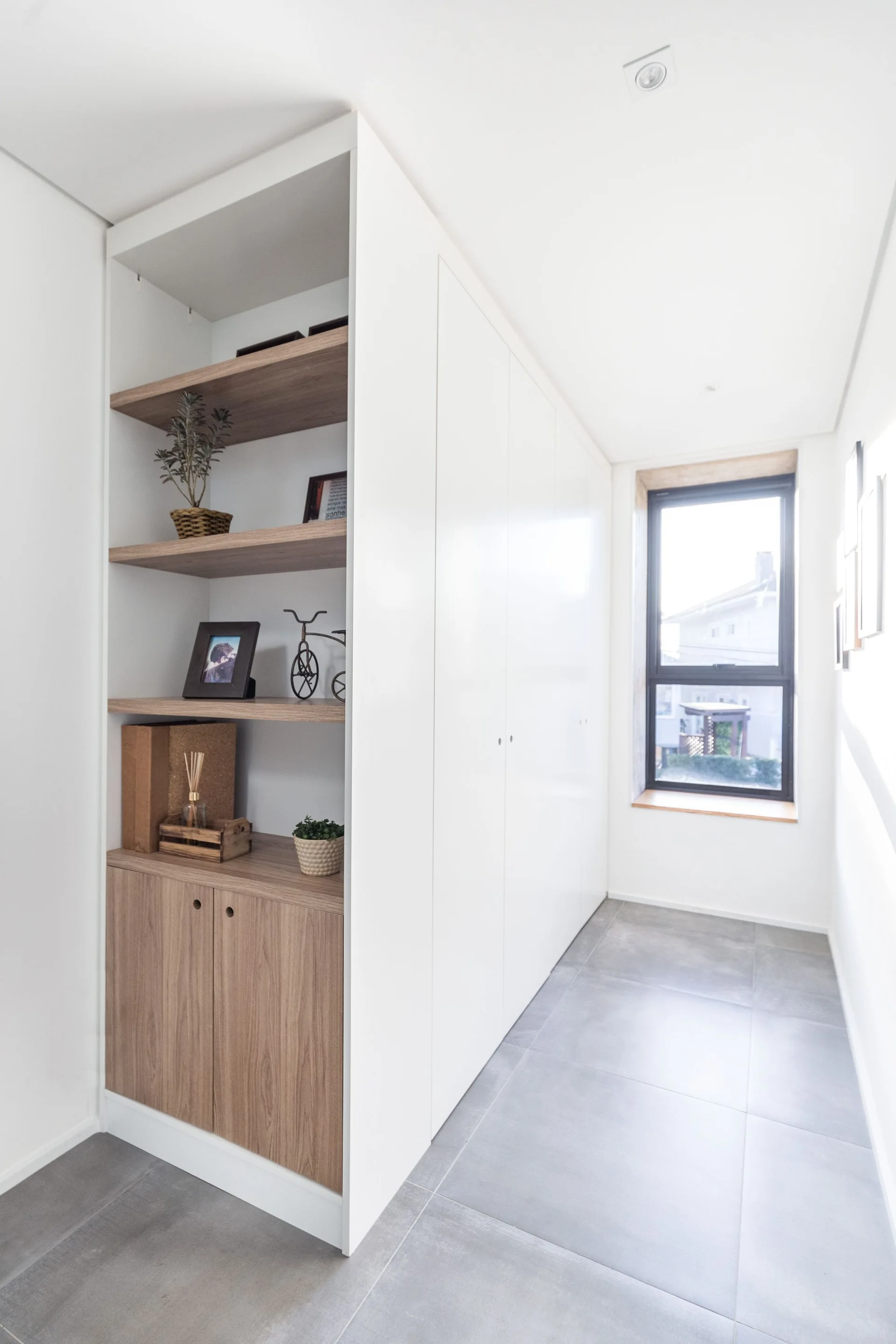 A bright hallway features a large white built-in wardrobe with open wood shelves on the side, grey tile flooring, and a window at the end.