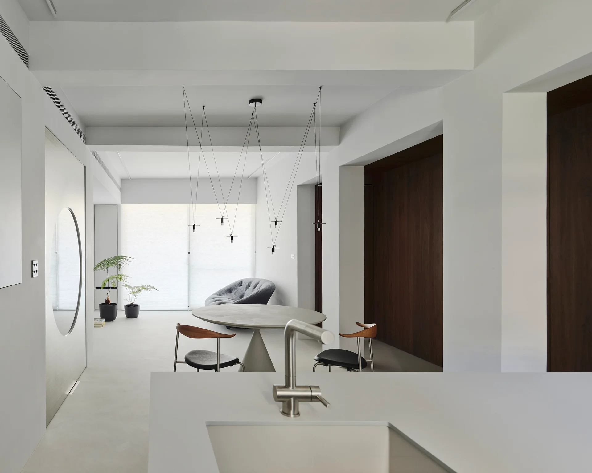 A minimalist open-plan kitchen and dining area features a round concrete-style table, thin black wire pendant lights, and seamless light gray floors.