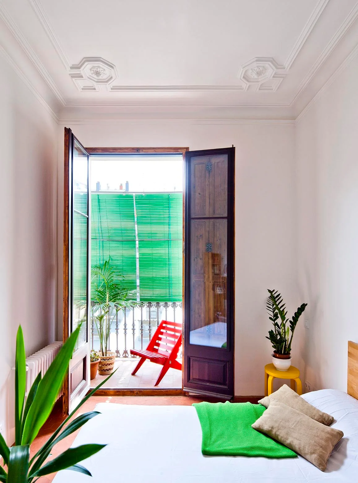 A bright bedroom with decorative white ceiling molding features French doors that open to a balcony with a red wooden chair.