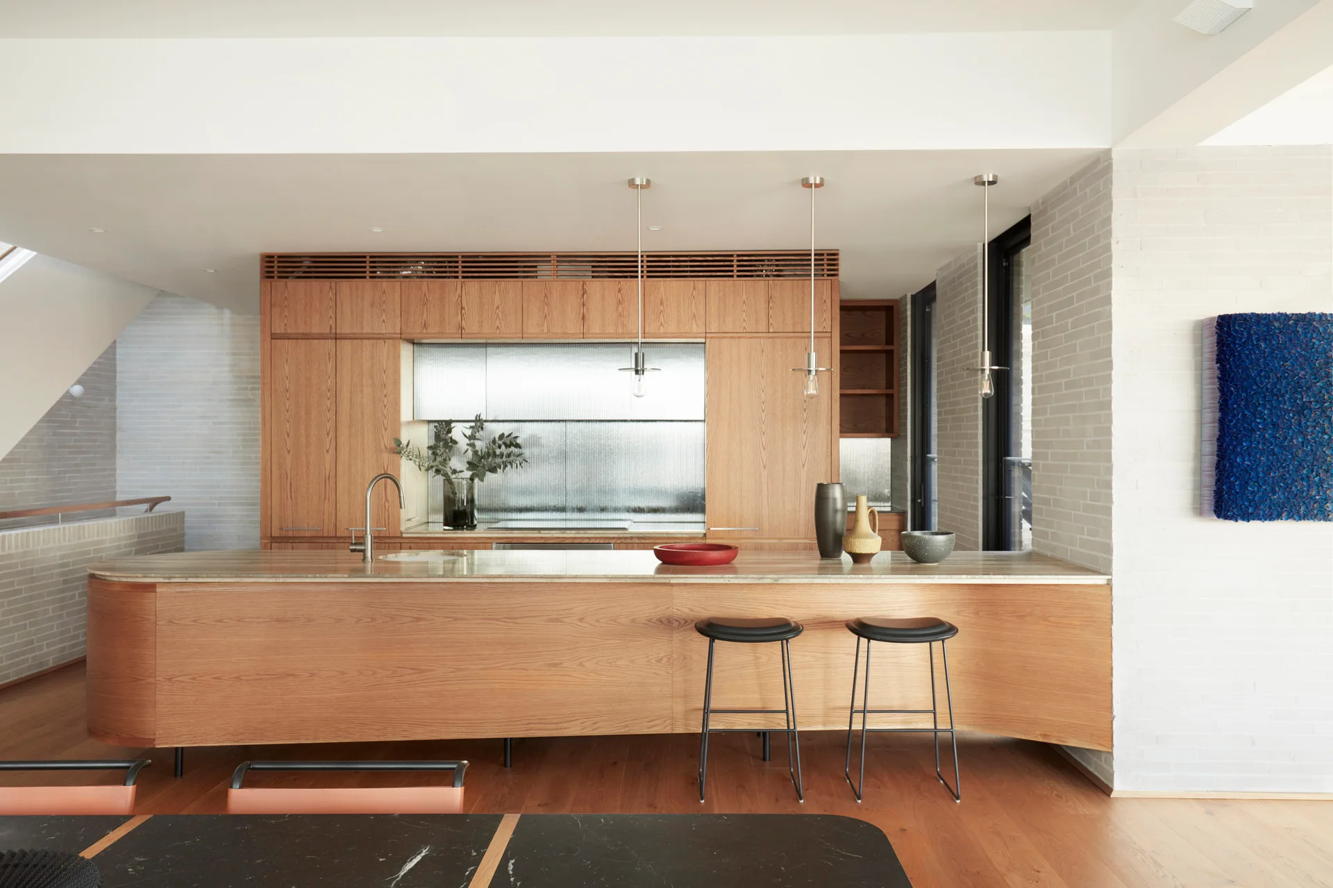 A modern kitchen features a curved wood island with two black bar stools, extensive flat-panel cabinetry, and a fluted glass backsplash.