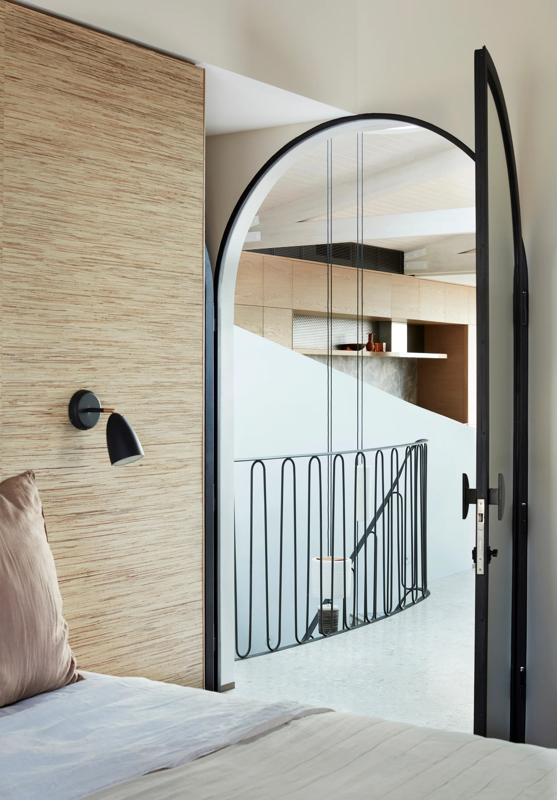 A bedroom view through a black steel-framed arched doorway leading to a hallway with a curved black metal stair railing.