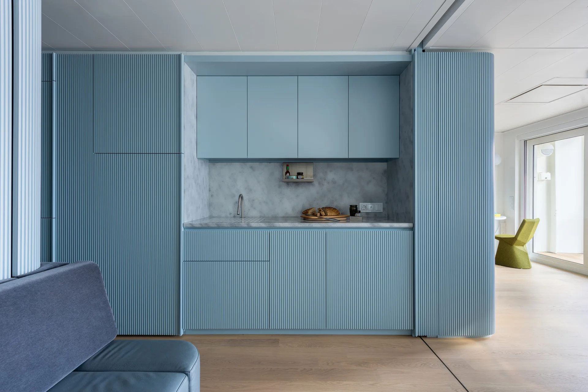 A minimalist kitchen featuring blue ribbed cabinetry, a grey stone countertop and backsplash with an integrated sink, and light wood plank flooring.