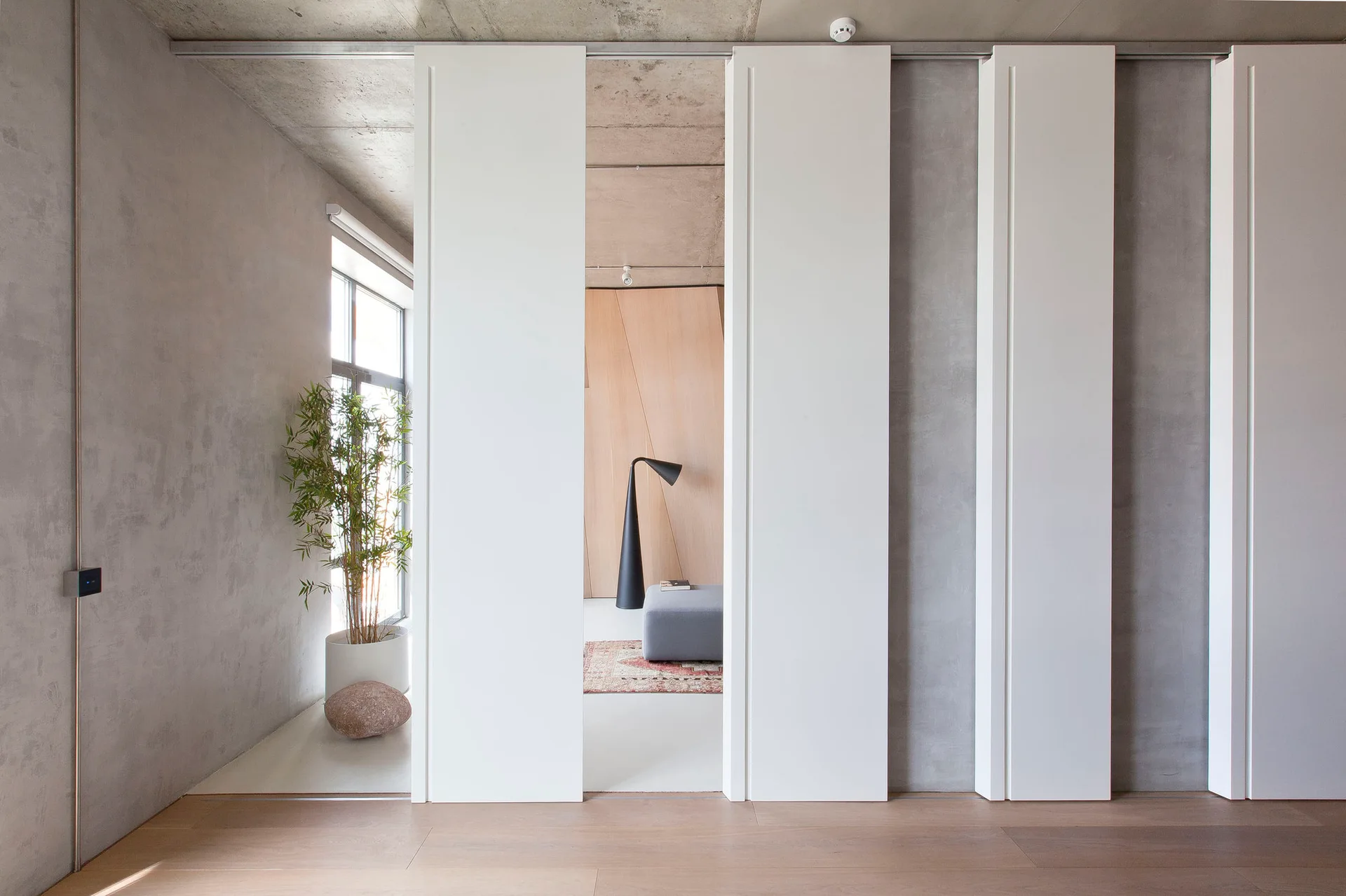 Large white sliding panels partition a modern room with raw concrete walls, light wood flooring, and a potted bamboo plant.