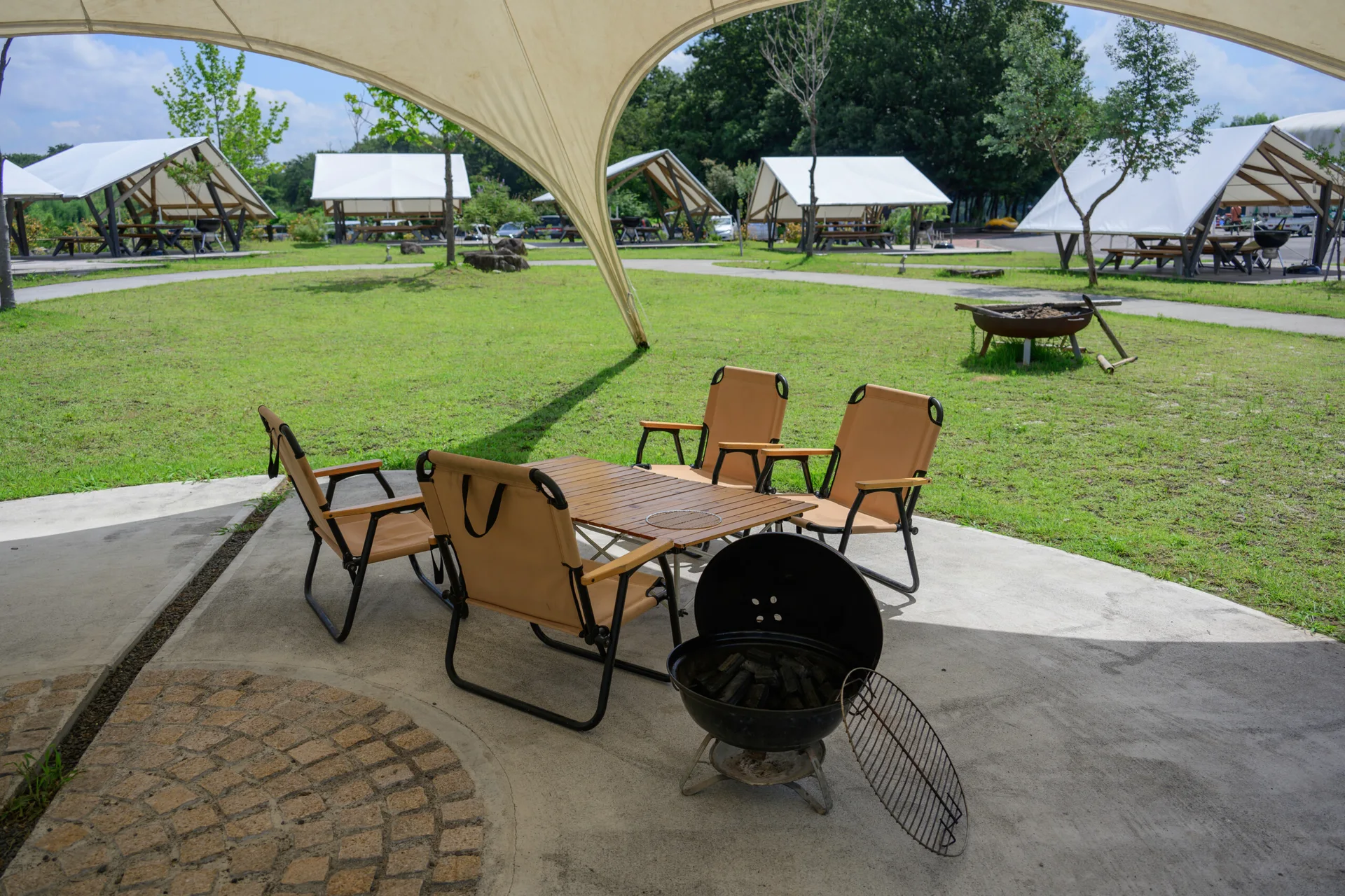Tan canvas folding chairs and a wood slat table sit on a concrete patio under a large white tensile canopy next to a black grill.