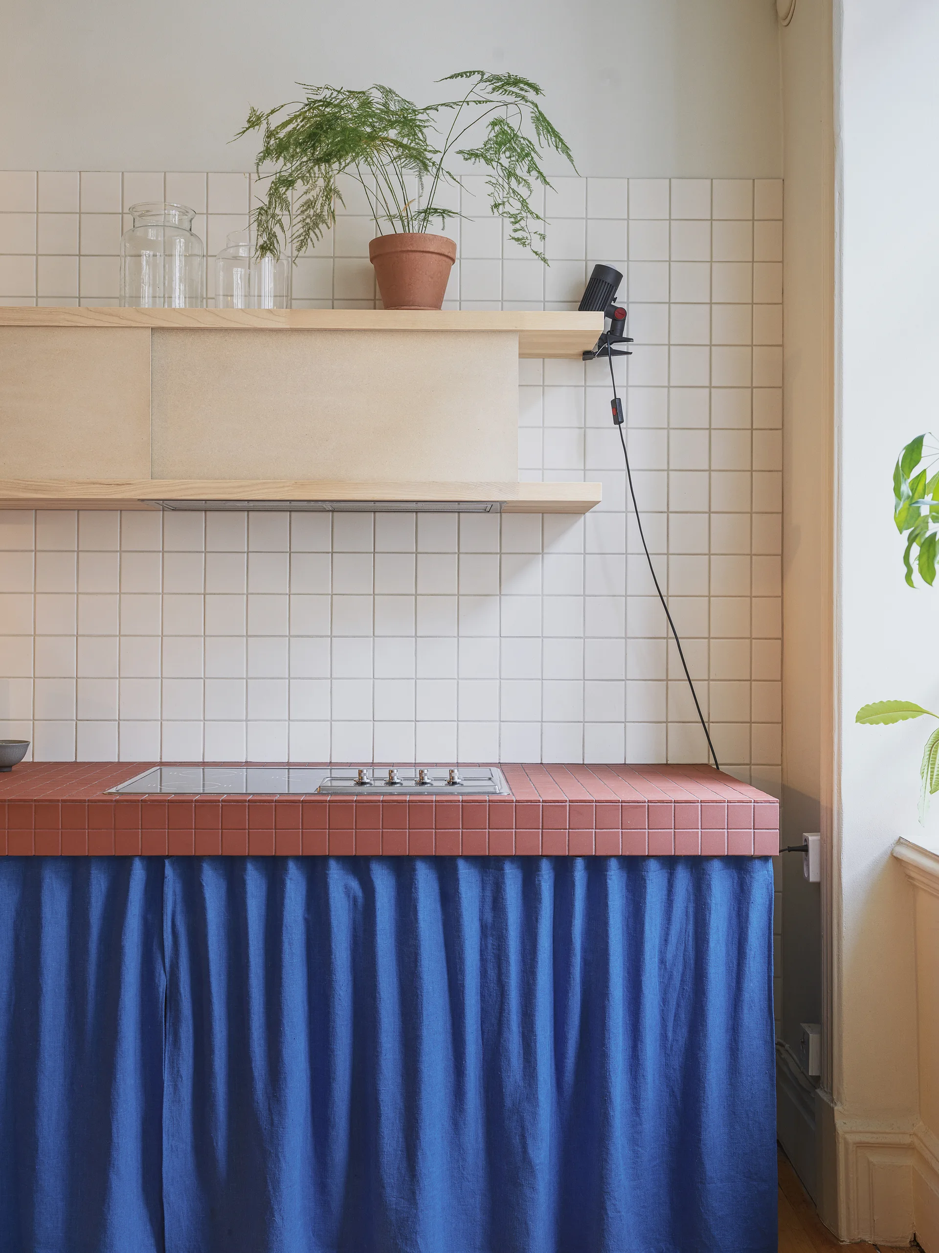 A kitchen counter featuring a terracotta-colored tile surface with a bright blue fabric skirt below and a white grid tile backsplash.
