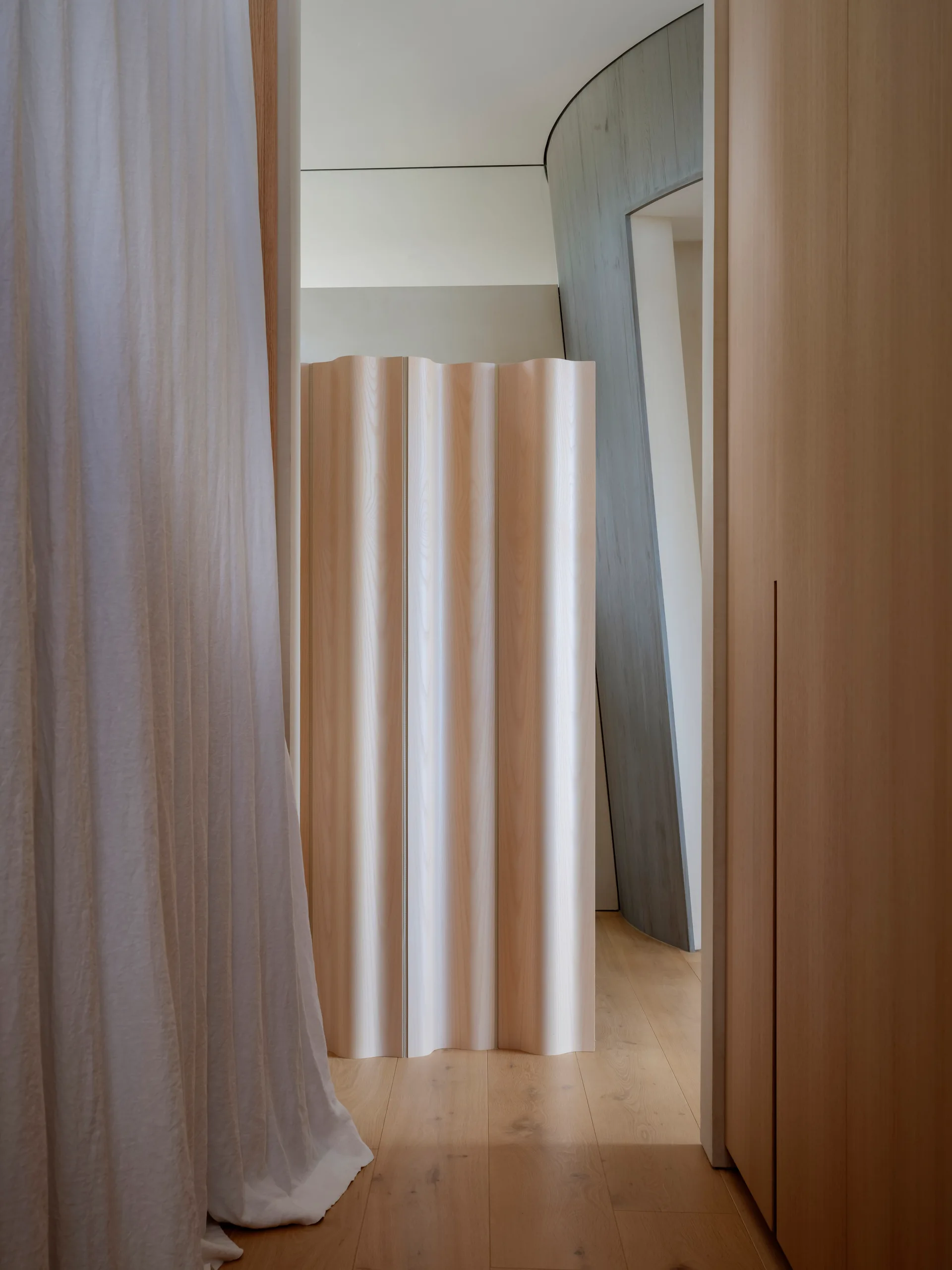 An undulating light wood room divider stands in a doorway between a white curtain and a wooden wall panel.