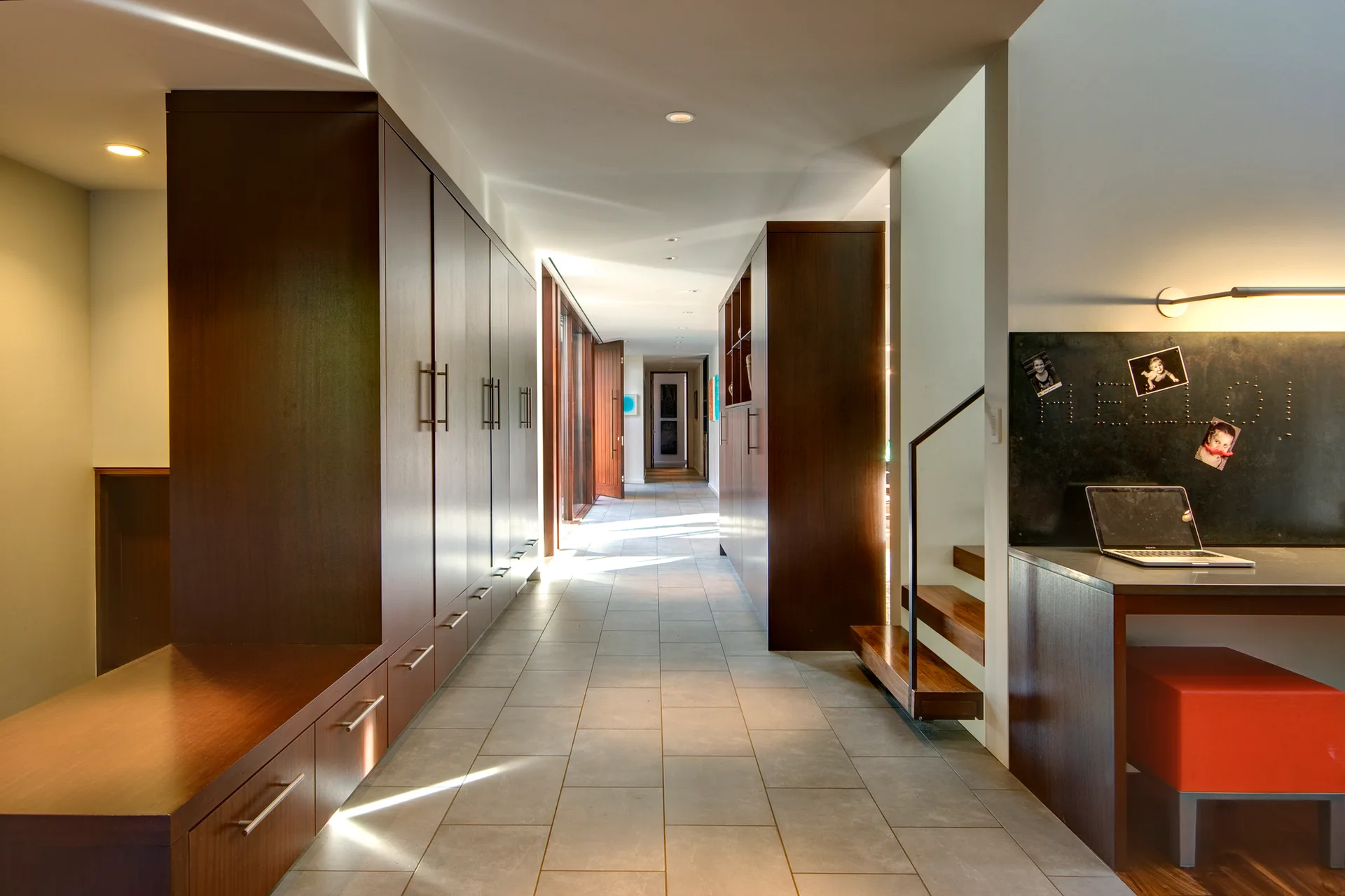 A long hallway features custom dark wood storage cabinets on the left, a built-in desk with a red-orange stool on the right, and large grey floor tiles.