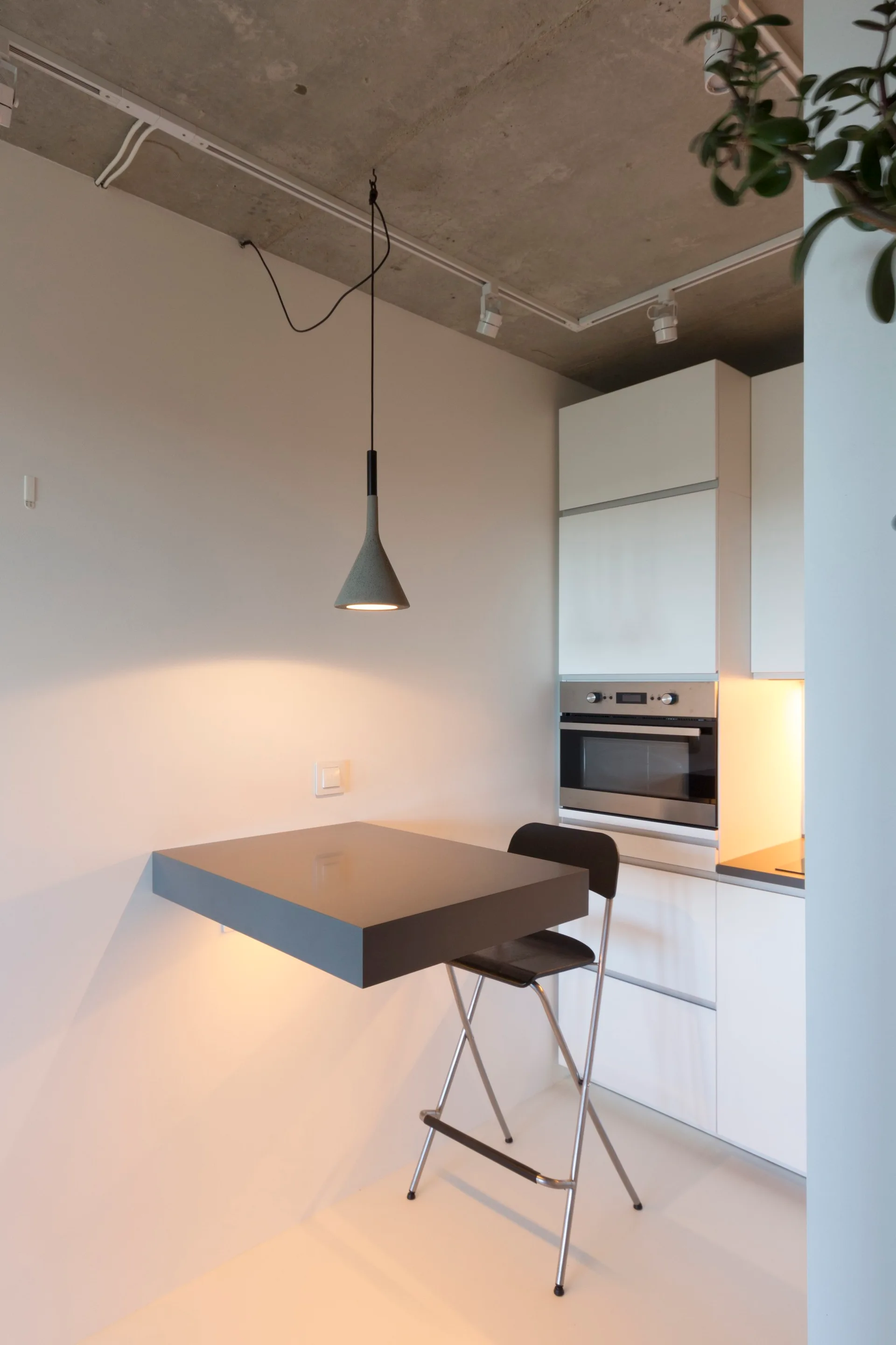 A minimalist white kitchen featuring a wall-mounted dining table, a black folding stool, and an exposed raw concrete ceiling.